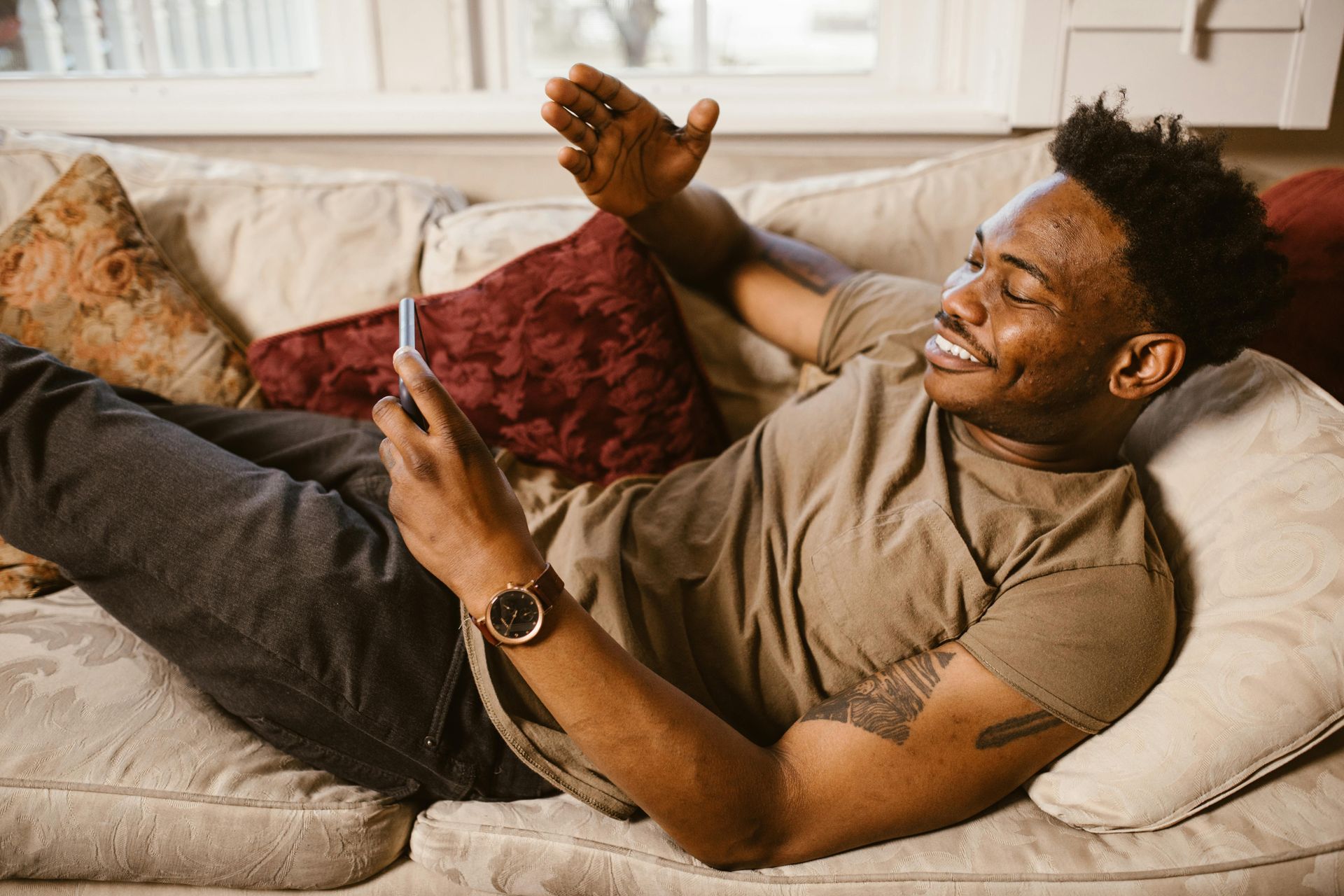 Man reclines on couch, smiles while looking at phone, and raises his hand.