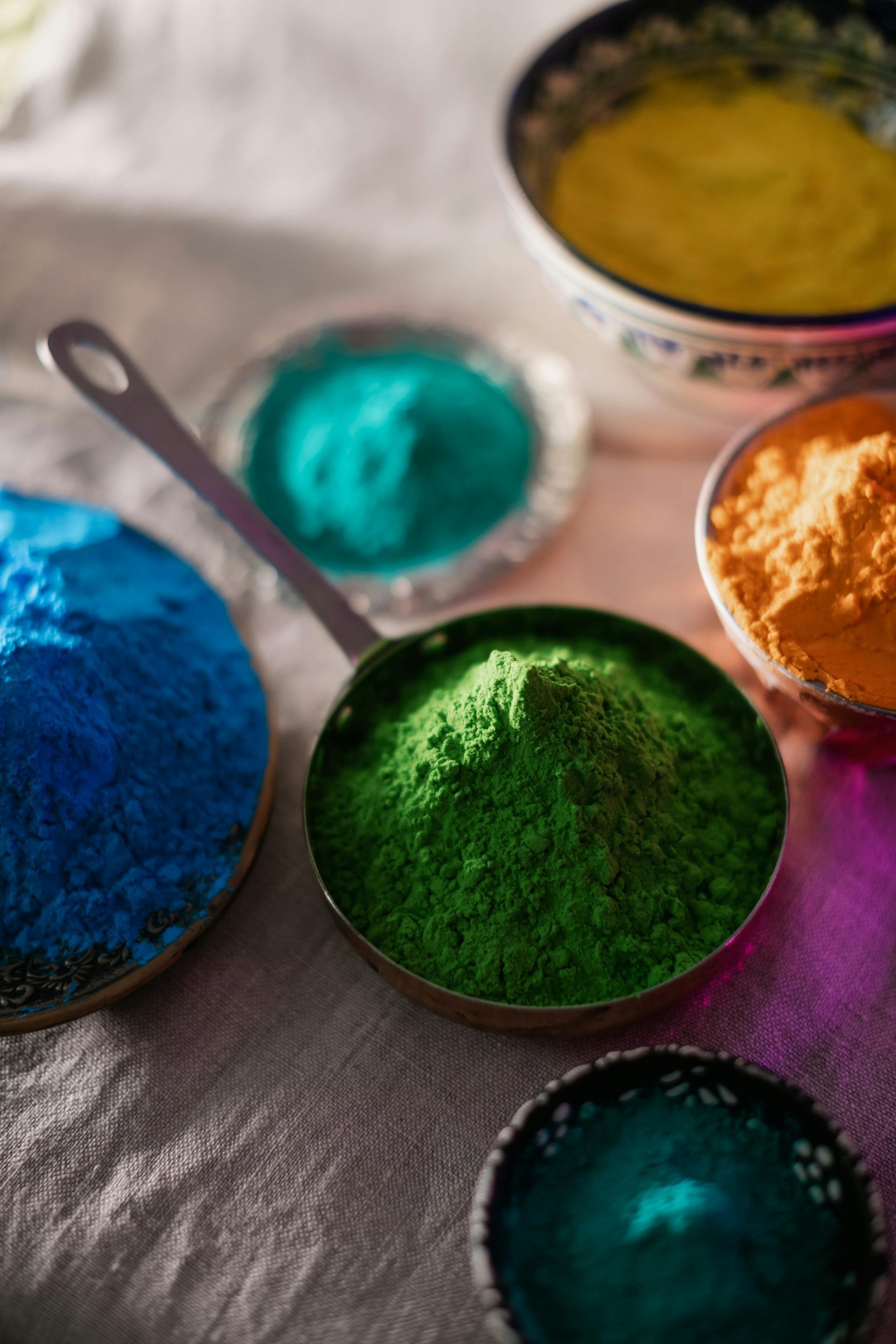 Bowls of brightly colored powdered pigment on a white cloth: blue, green, teal, orange, and yellow.