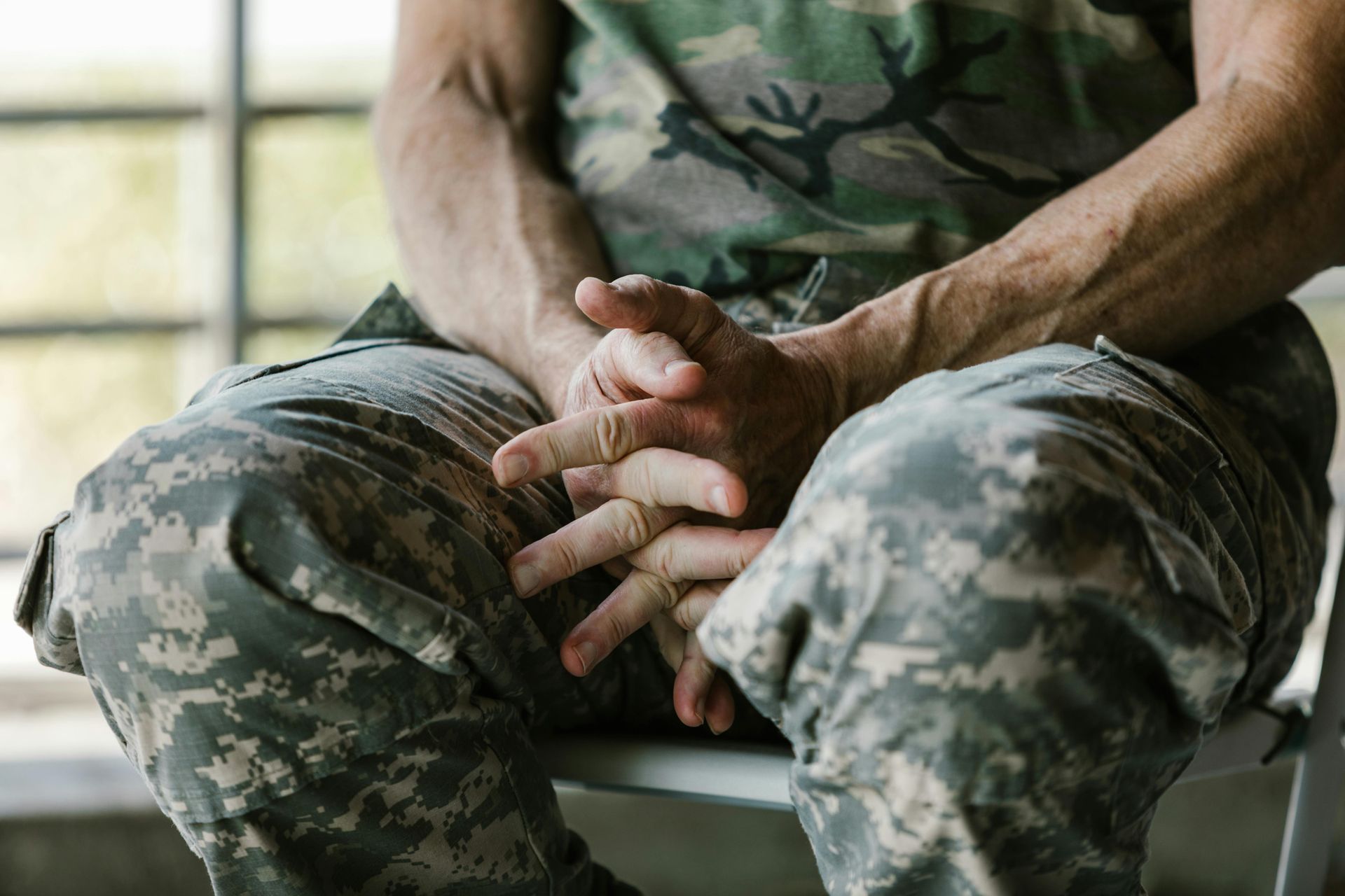 A person in camouflage uniform sits with hands clasped.