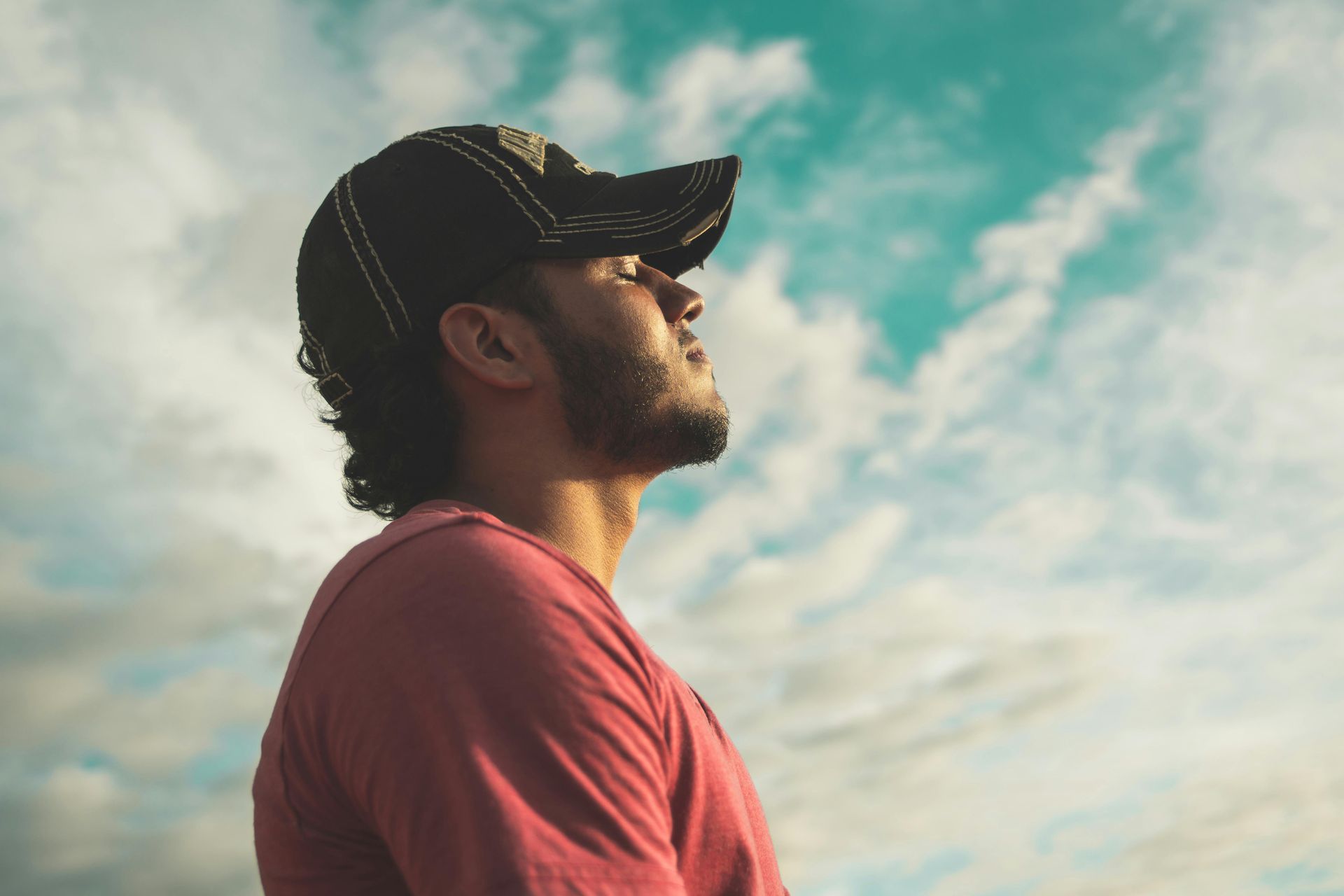 Man wearing a cap looking up with eyes closed against a cloudy sky.