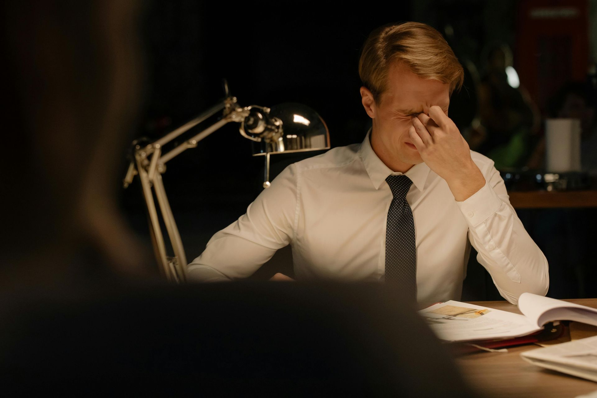 Man in white shirt and tie at desk, rubbing his eyes, looking stressed. Desk lamp on.