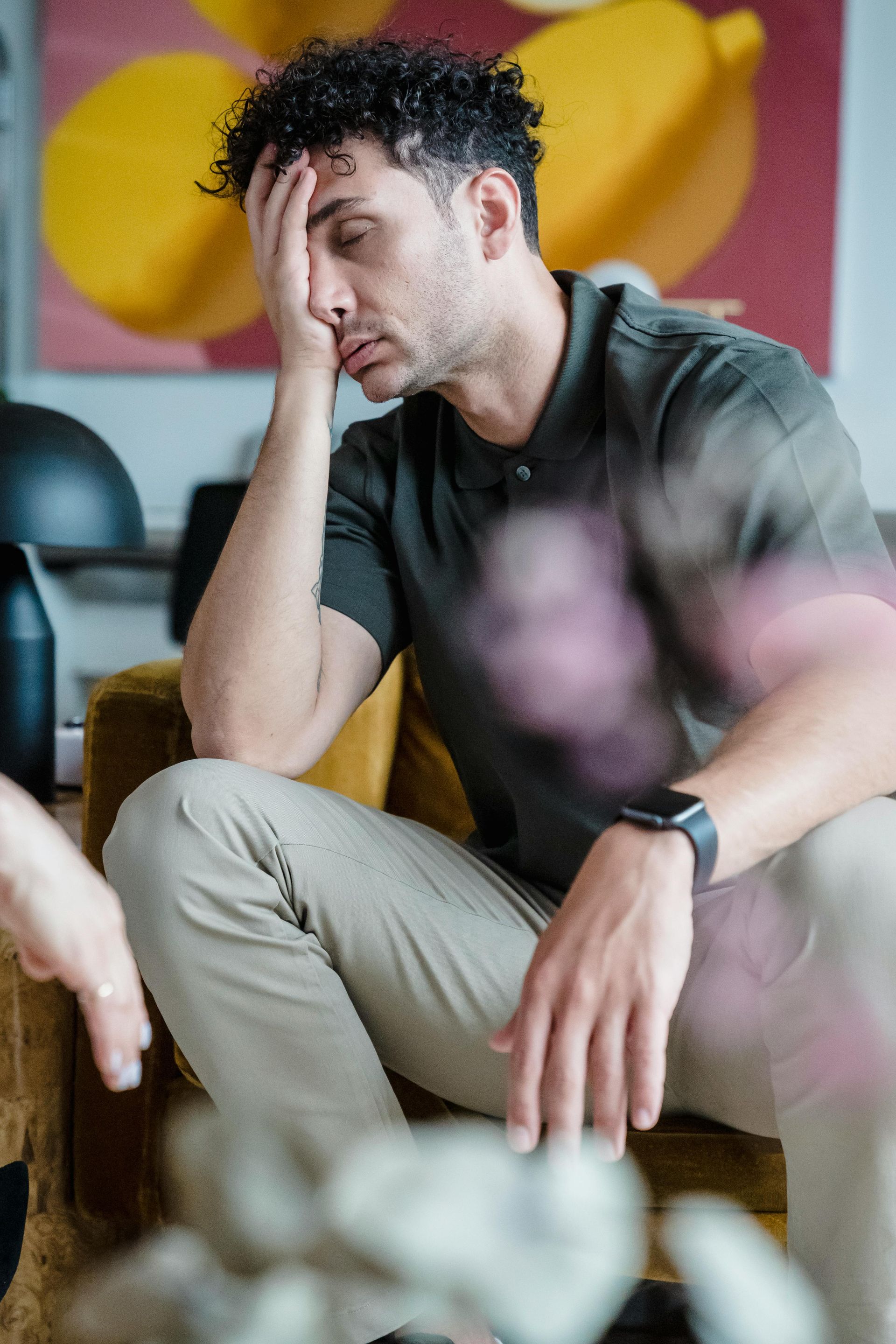 Man in a green shirt and khaki pants, resting his head. He sits on a yellow chair, looking tired.