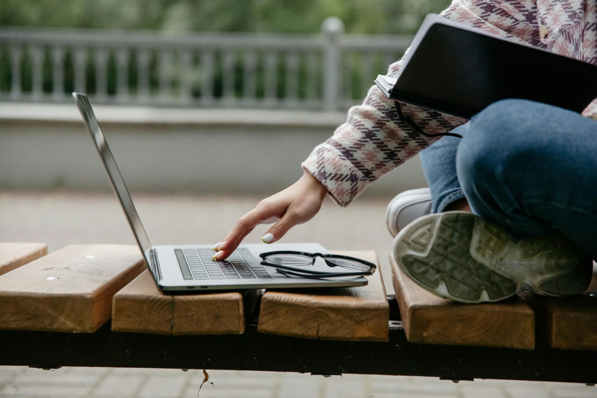 Person using laptop on a wooden bench, with eyeglasses, and a notebook in hand, outdoors.