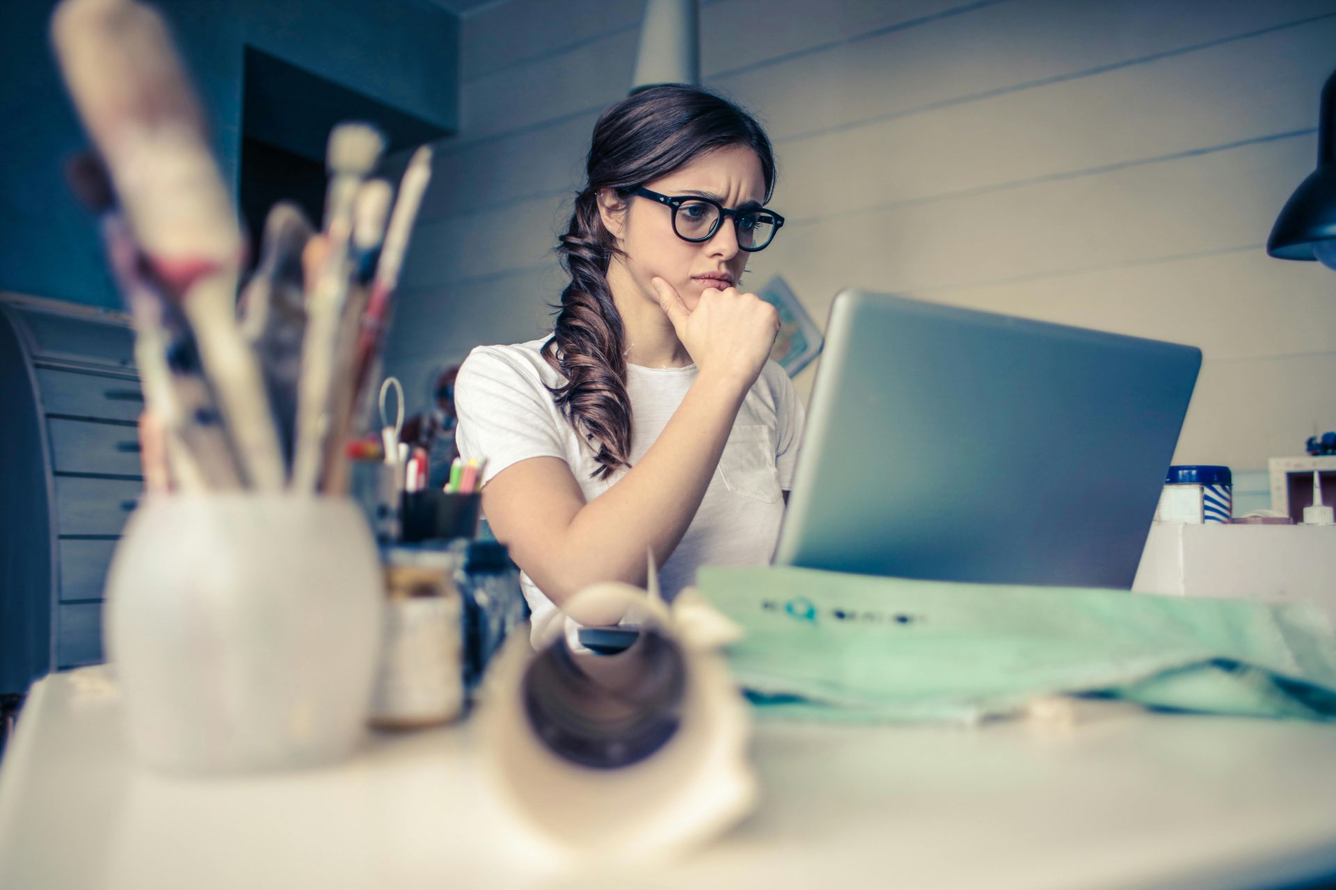 Woman with glasses, working on a laptop, looking thoughtful with her hand on her chin, with art supplies on a desk.
