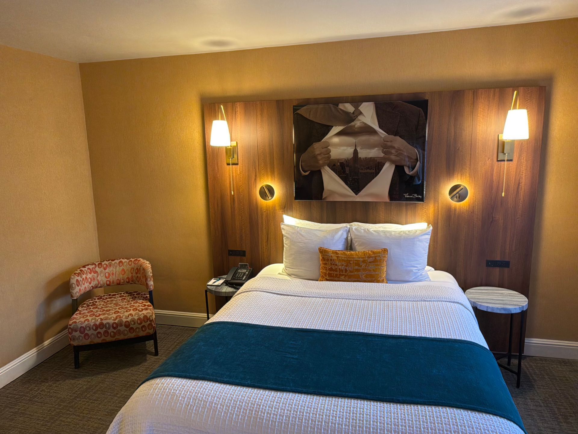 A hotel room with a queen bed featuring a blue runner, a patterned armchair, bedside tables, and wall art above the bed.