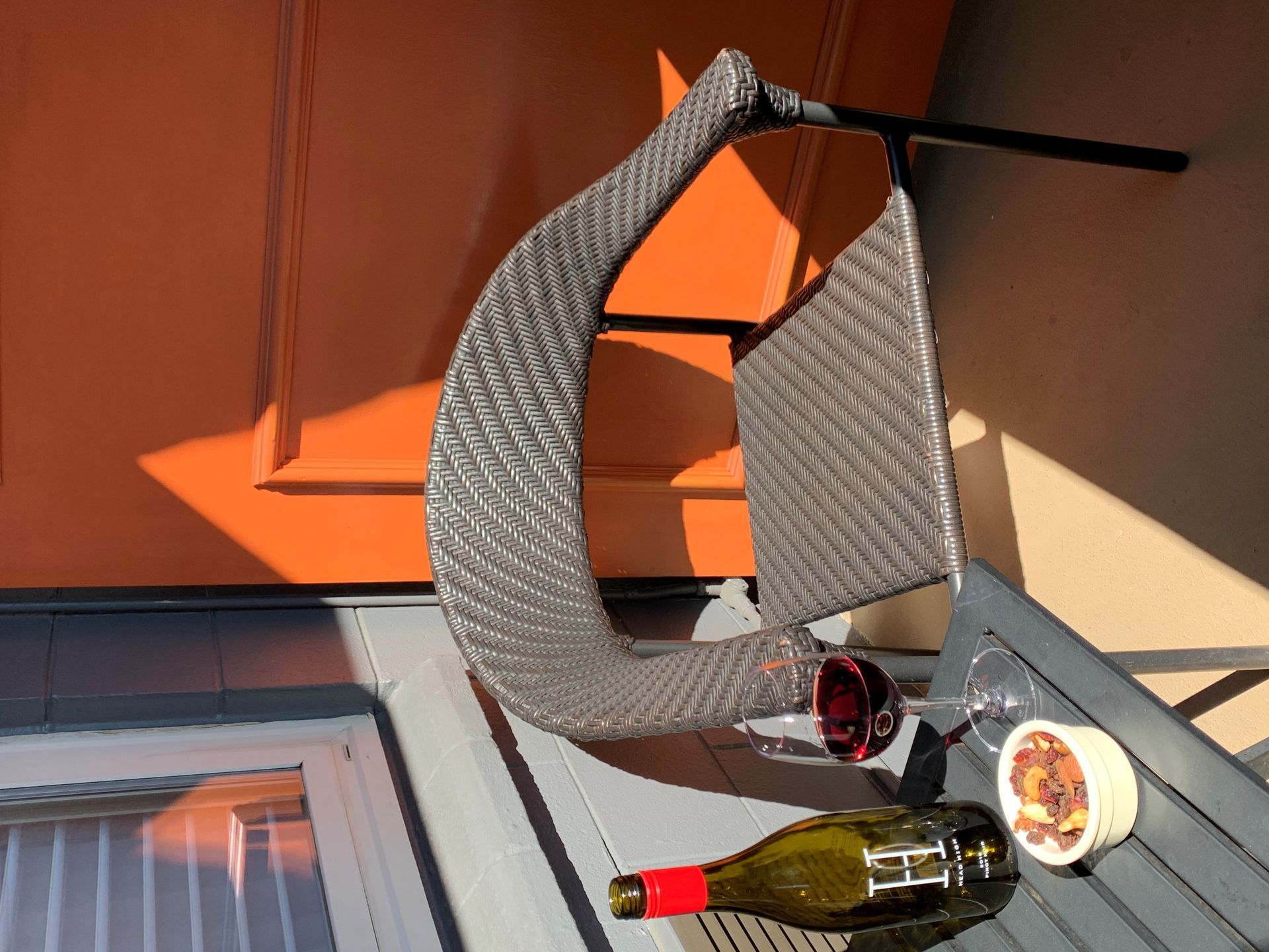 A wicker chair, a glass of red wine, and a bottle on a metal table set against an orange wall in bright sunlight.