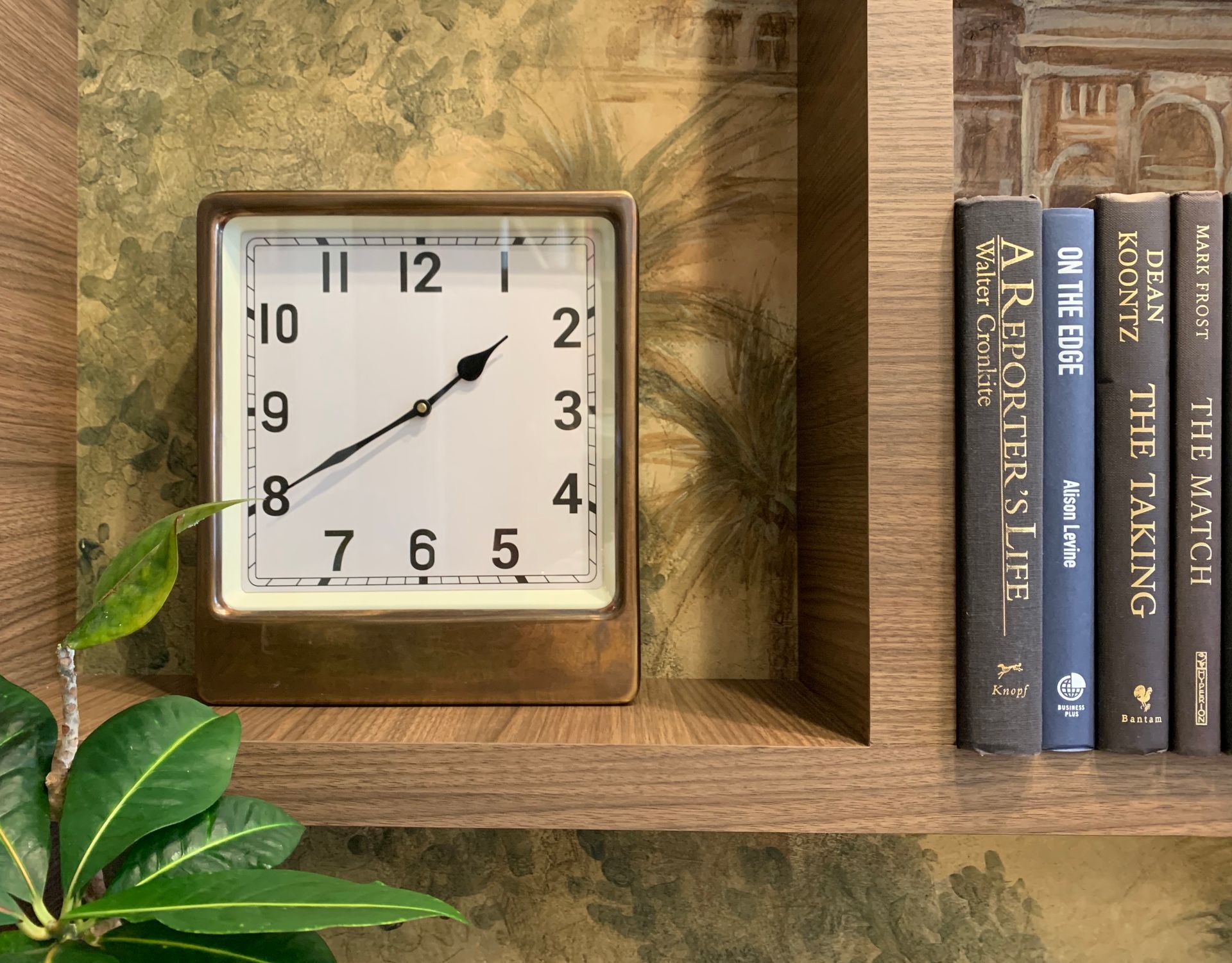 A square, brass-framed clock sits on a wooden shelf next to a row of books, with decorative wall wallpaper in the background.