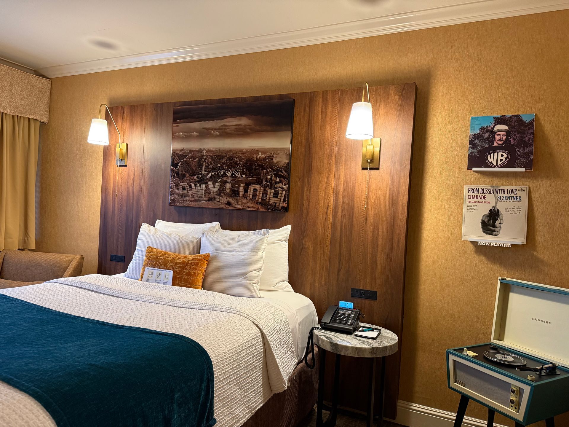 A hotel room featuring a bed with white bedding, a teal throw, and a wooden headboard with two wall lamps and art.