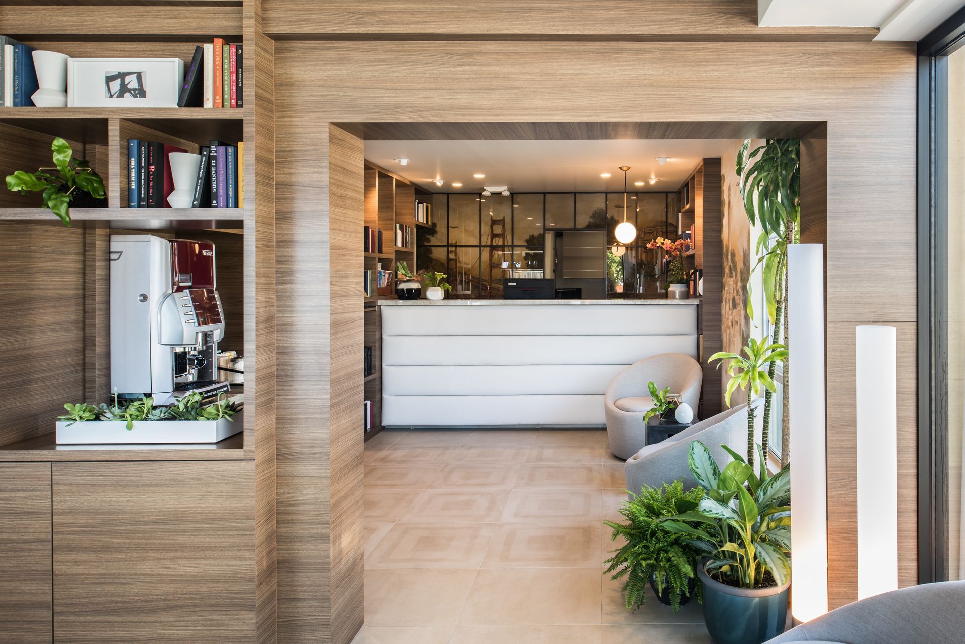 A view into a modern hotel lobby with wood-paneled walls, a white front desk, cozy armchairs, and lush indoor plants.