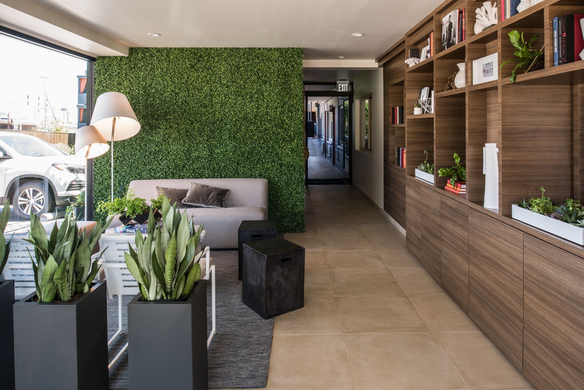 Modern waiting area with a green wall, sofa, tall potted plants, floor lamp, and a large wooden bookshelf.