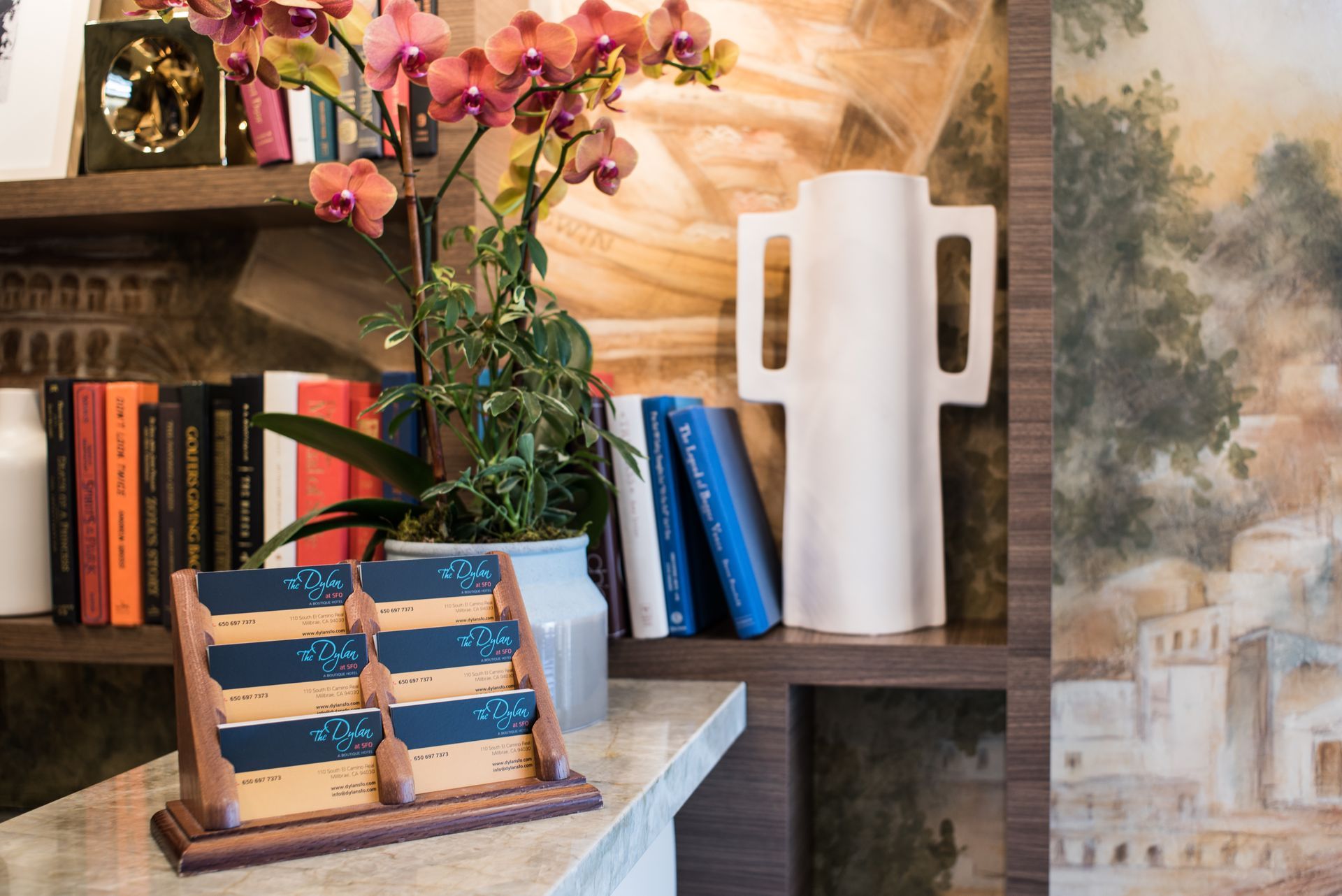 A desk display holder with cards sits on a counter next to a potted orchid and a white vase on a shelf of books.