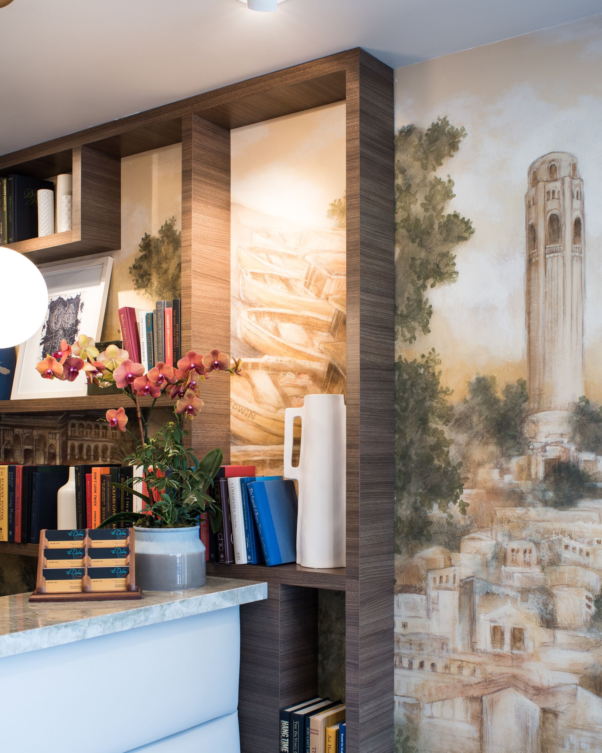 A wooden shelving unit with books and orchids sits against a wall featuring a mural of Coit Tower in San Francisco.