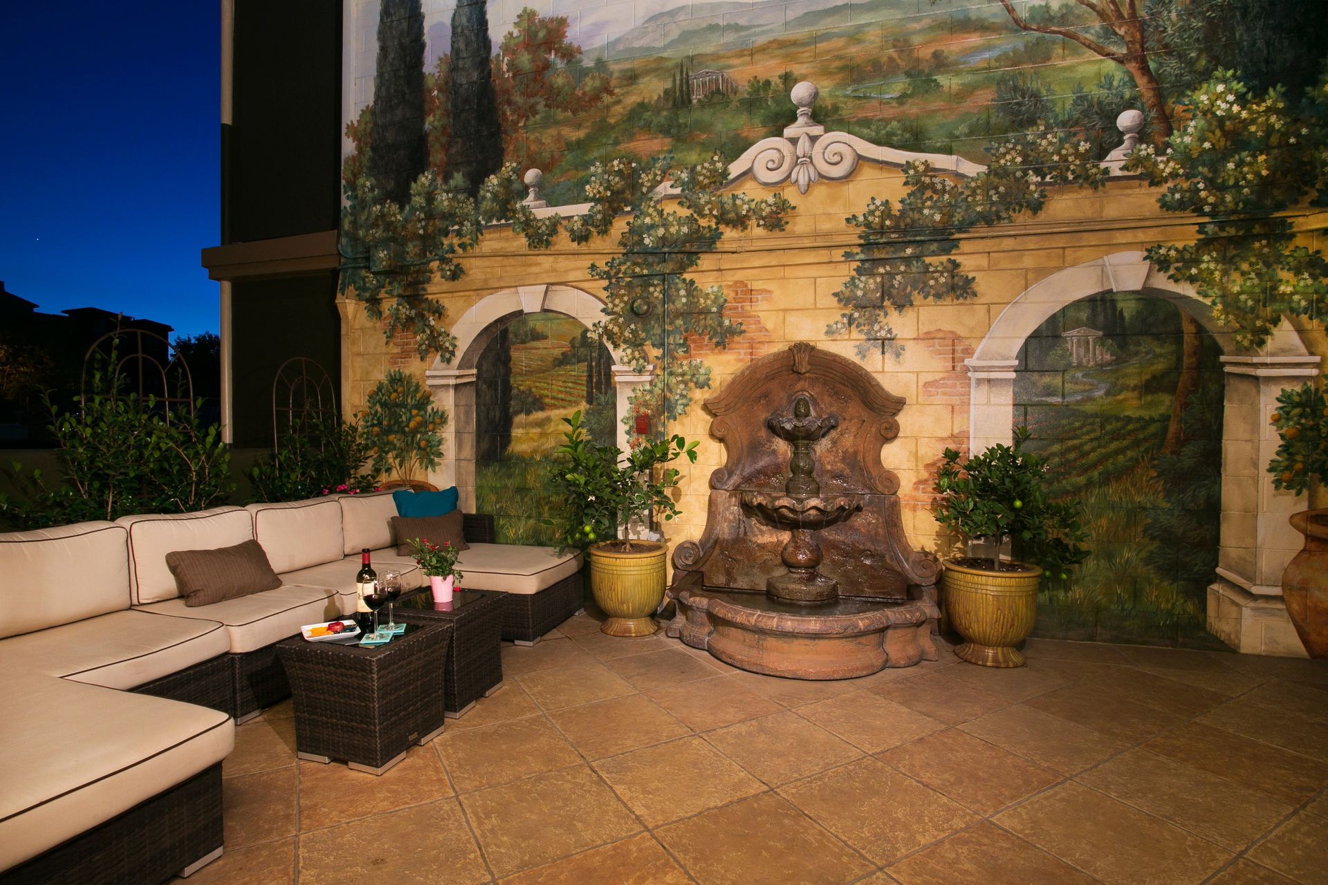 An outdoor patio at dusk with beige cushioned lounge seating, a fountain, and a large mural of an Italian landscape.
