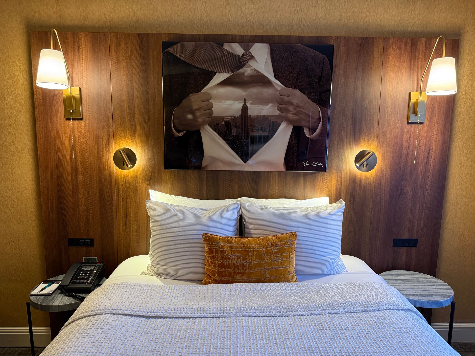 A hotel bed with striped pillows, a gold accent pillow, and a wall-mounted artwork of a city inside an open shirt.