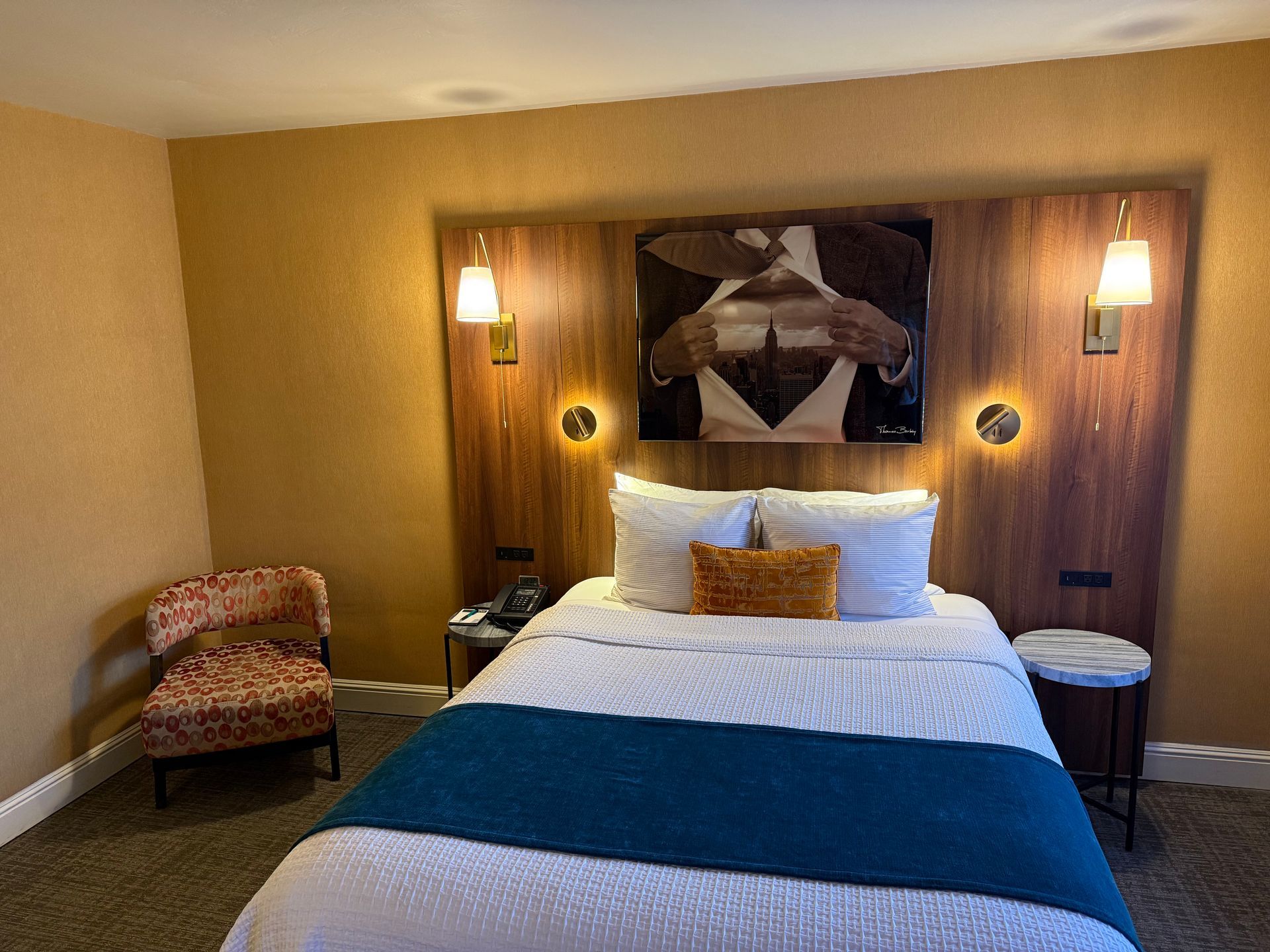 A hotel bedroom featuring a queen bed with a teal runner, tan walls, a framed wall art piece, and a patterned armchair.