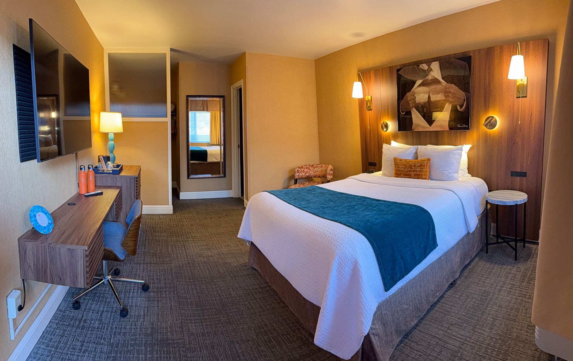 A hotel room with a queen bed featuring a blue runner, a wooden desk with a chair, a TV, and a decorative headboard.
