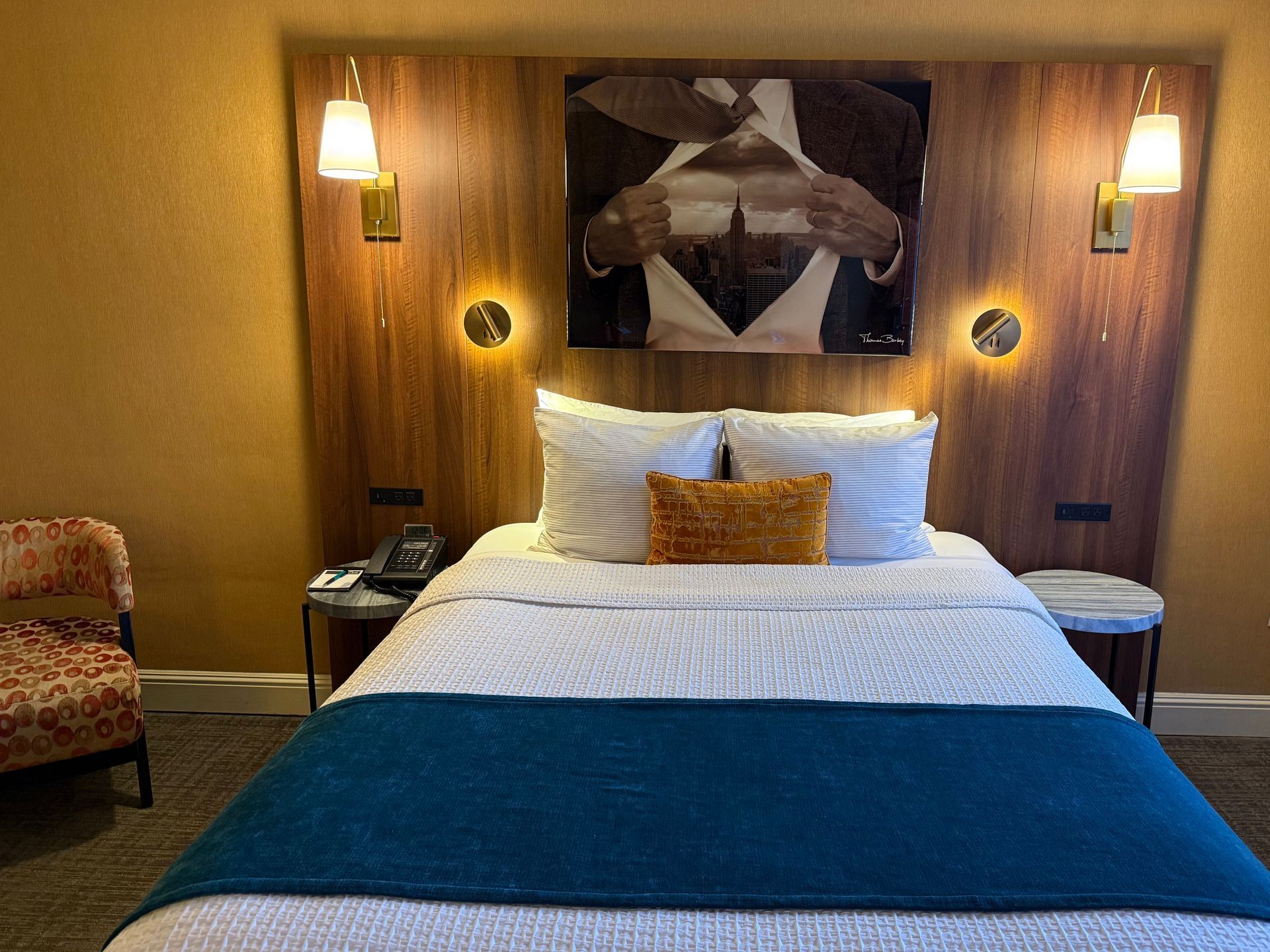 A bedroom featuring a queen bed with white linens, a teal runner, a golden pillow, and wall art above a wooden headboard.