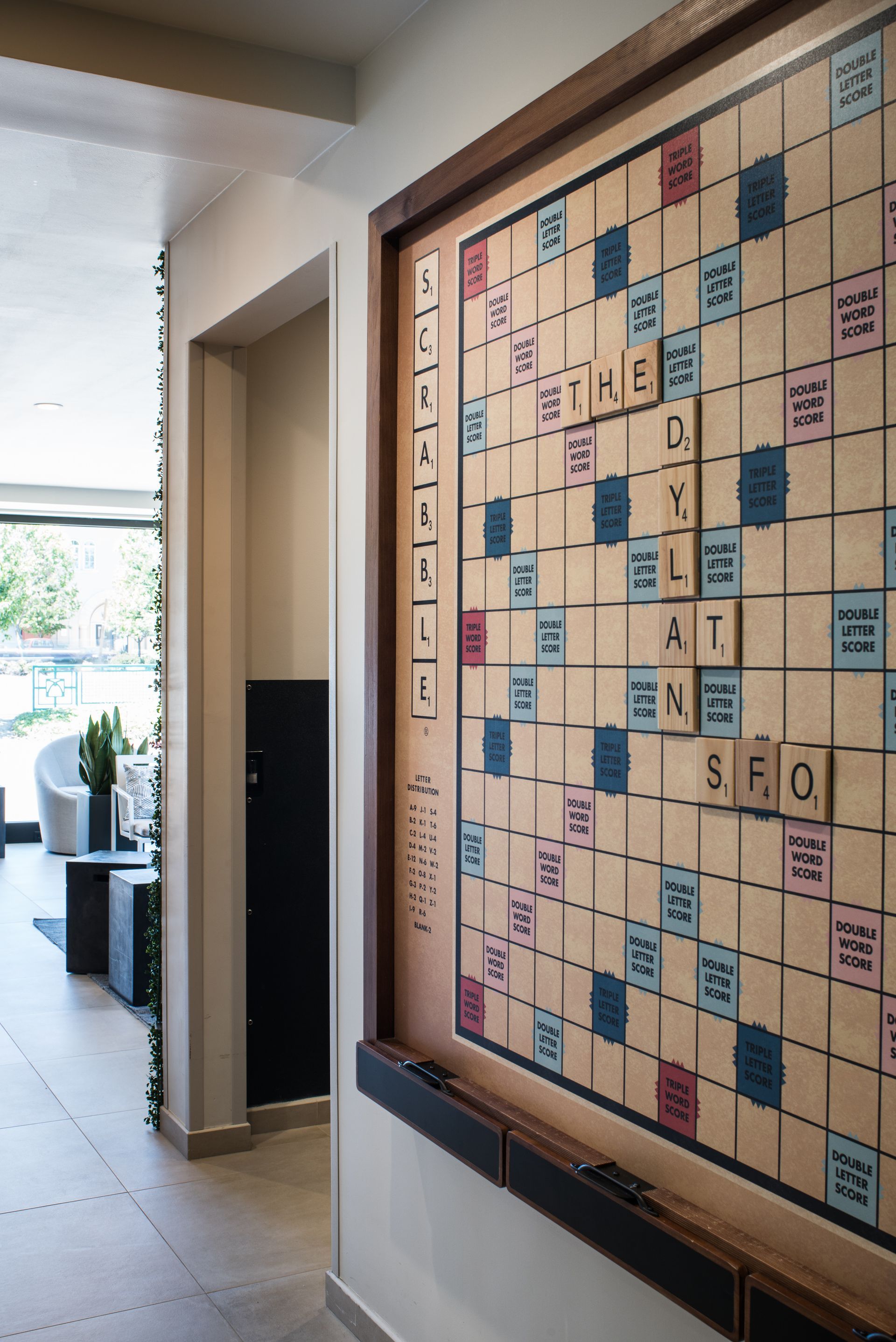 A large wall-mounted Scrabble board game hangs in a light-filled hallway, partially obscuring a doorway.