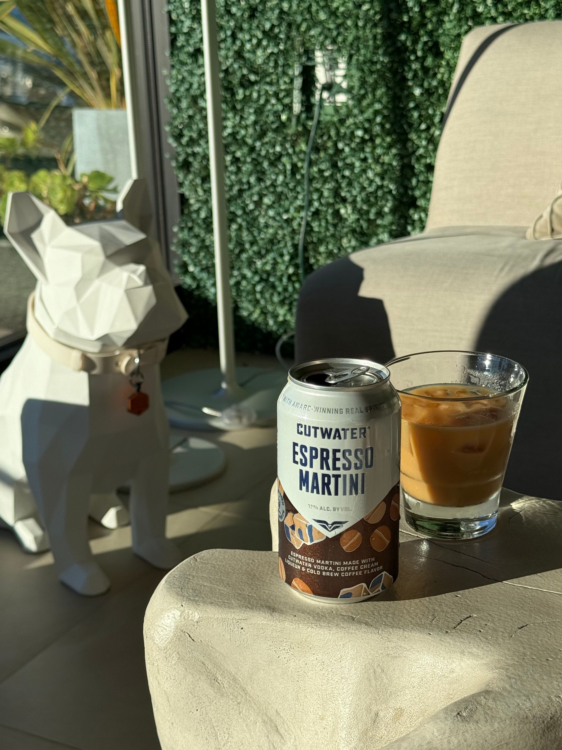 A can of Cutwater Espresso Martini beside a glass of the cocktail on a stone surface next to a geometric dog statue.