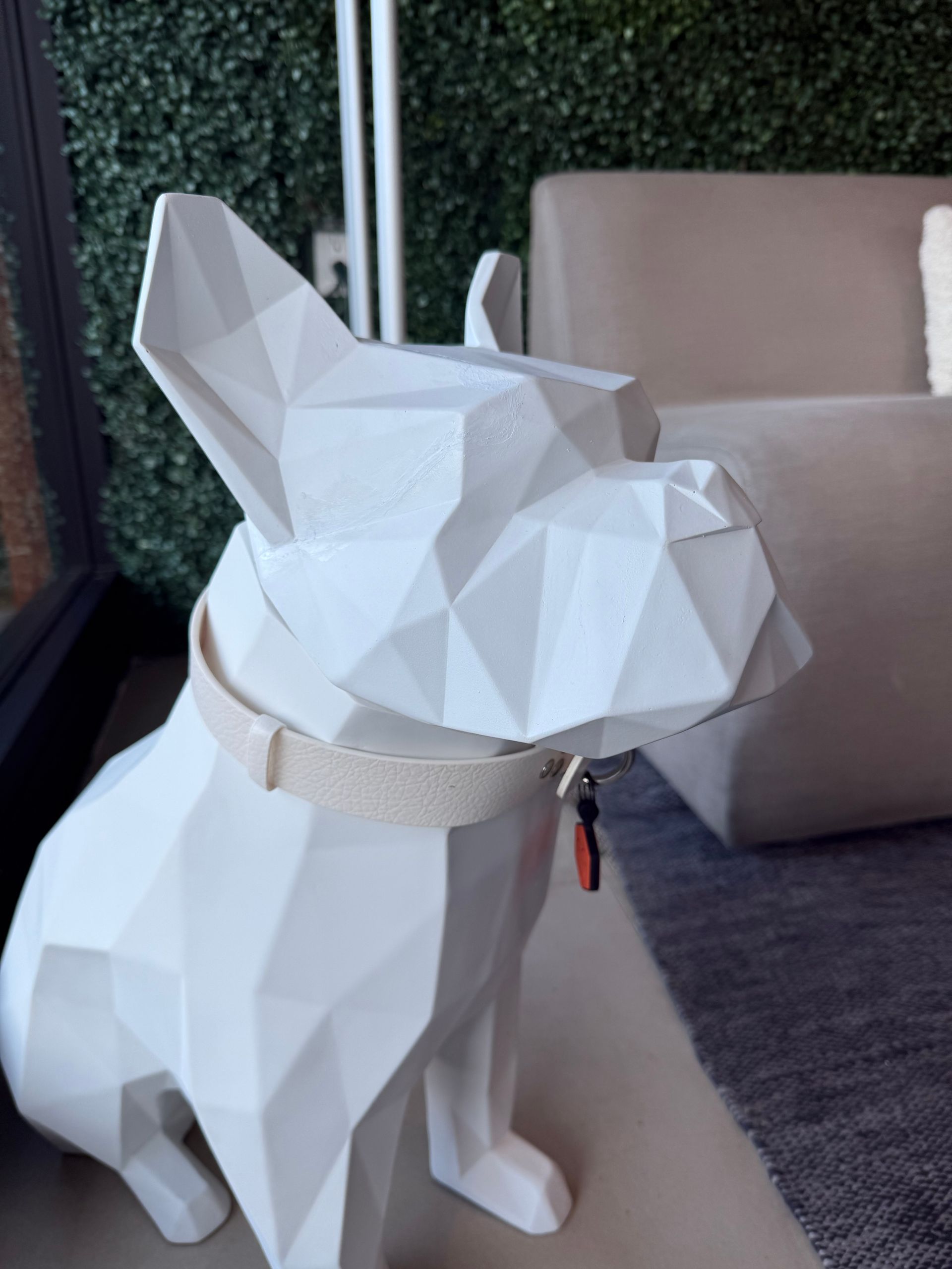 A white, low-poly geometric dog sculpture wearing a fabric collar with a small tag, sitting indoors near a sofa.