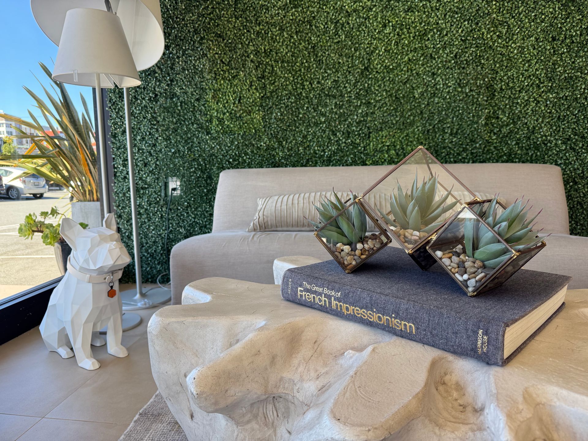 A white dog figurine, modern geometric planters on a book, and a beige sofa set against a green wall in a sunlit room.