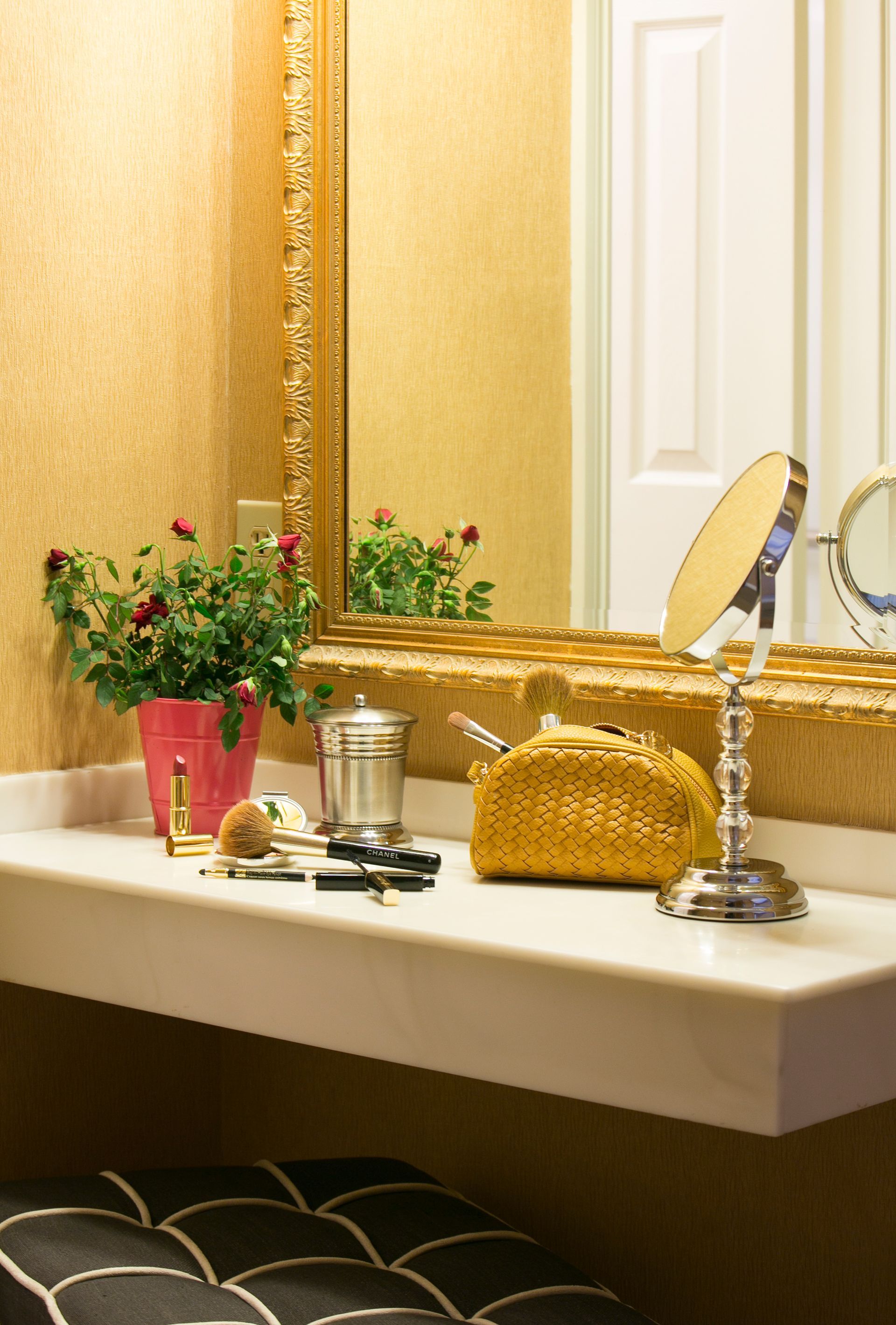 A vanity table with a gold-framed mirror, a rose plant in a pink pot, a gold pouch, and a magnifying makeup mirror.