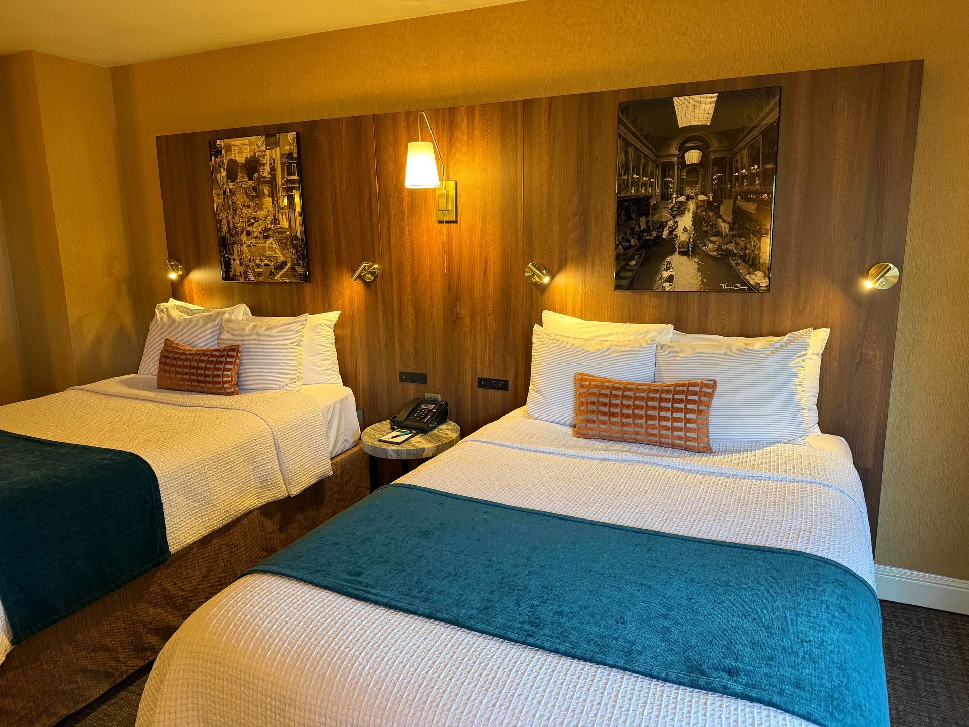 A hotel room featuring two beds with white patterned bedding and teal runners, set against a dark wood headboard wall.