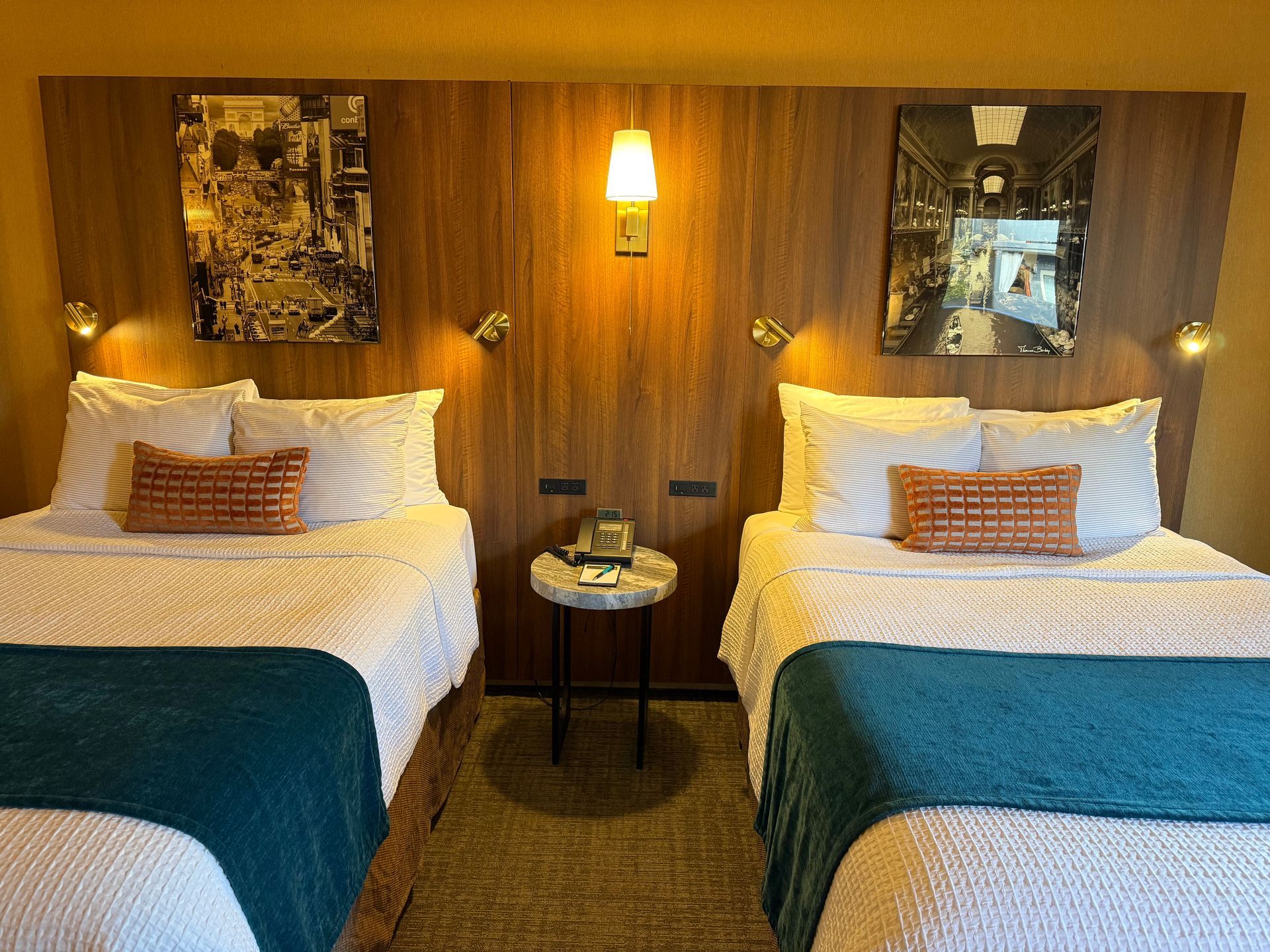 Two beds with white linens, blue runners, and patterned accent pillows against a wood-paneled wall with wall lamps.