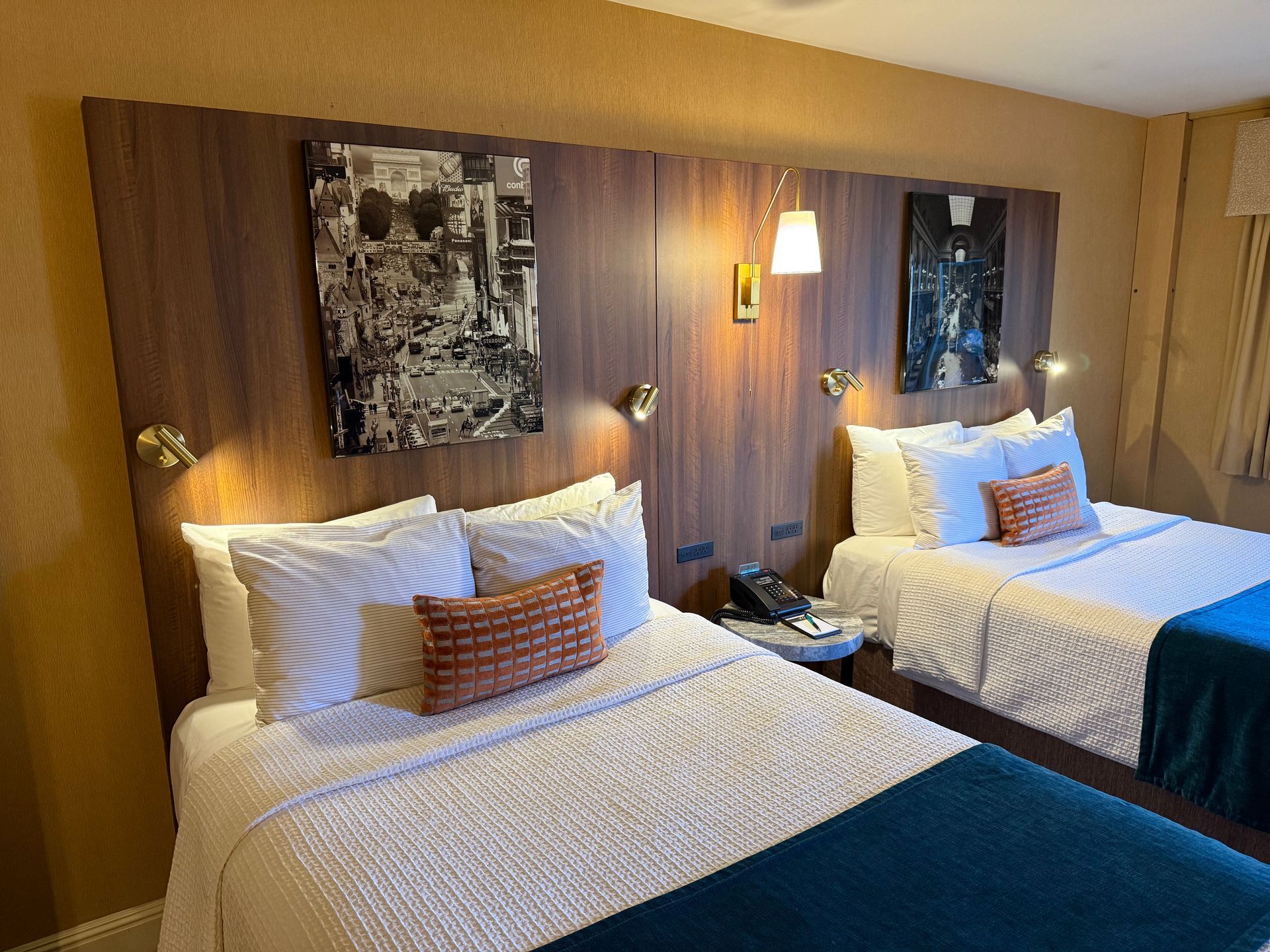 Two hotel beds with white patterned quilts, blue runners, and decorative orange pillows against a wooden headboard.