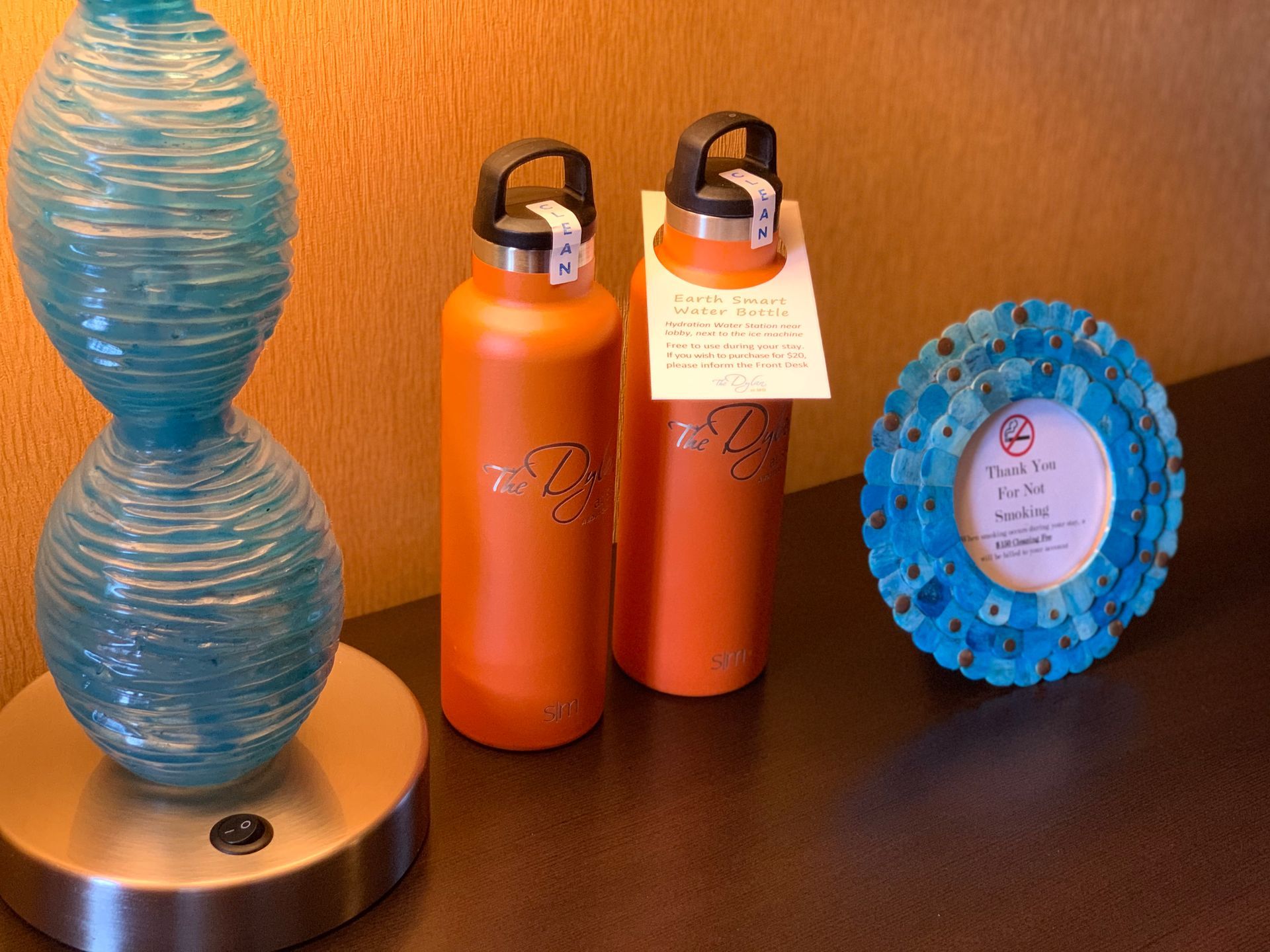 A blue textured lamp, two orange reusable water bottles, and a round, blue mosaic frame sit on a dark surface.