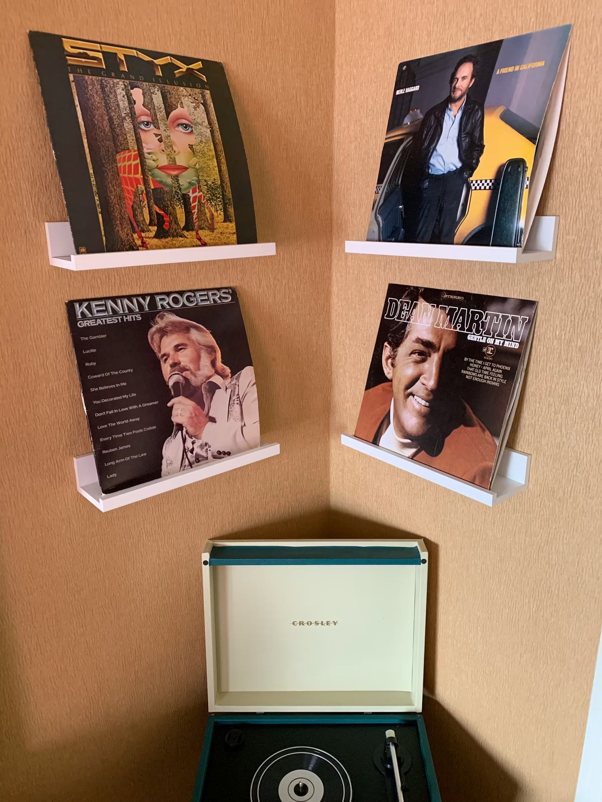 Four vinyl records are displayed on white corner shelves above a teal portable record player.