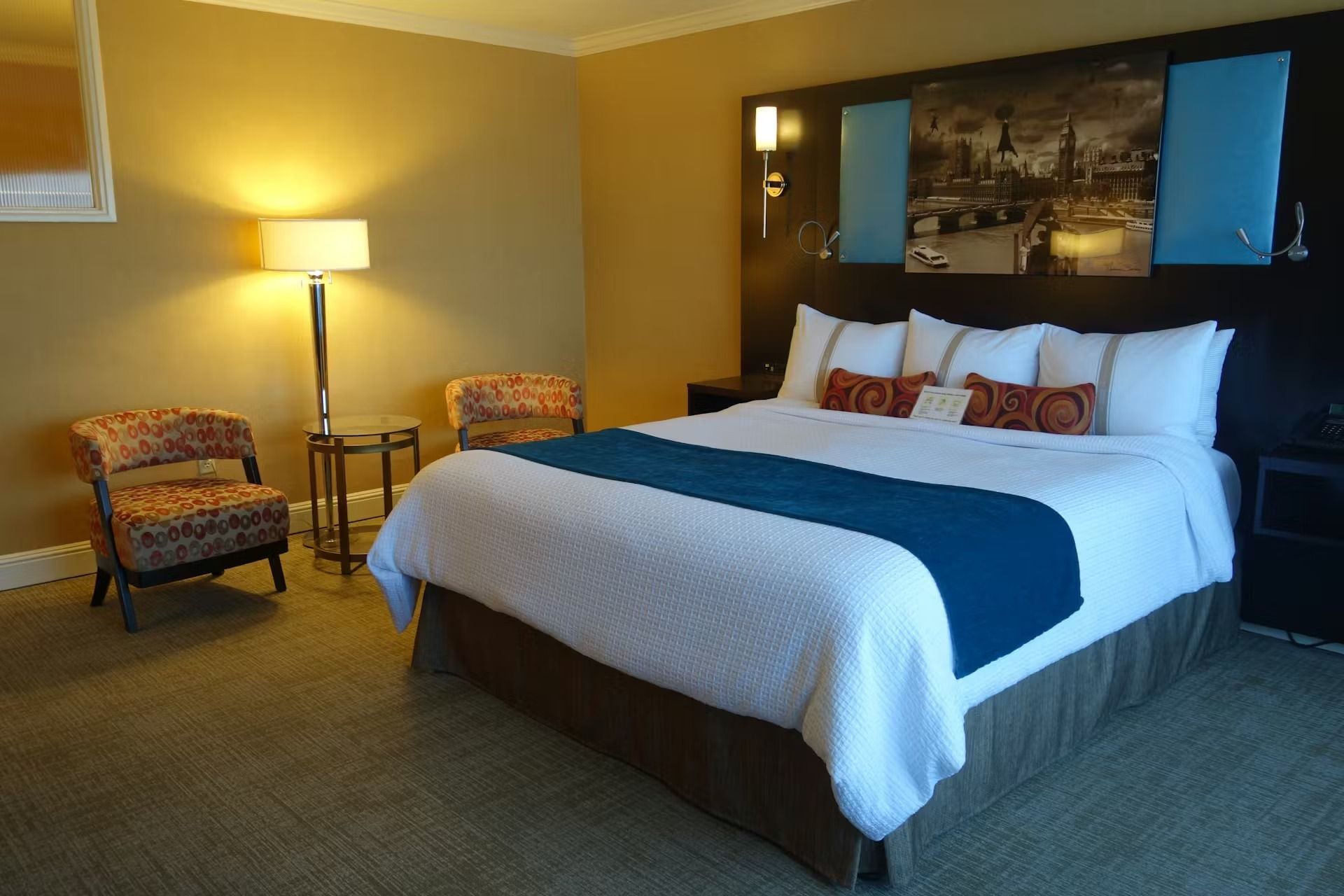 A modern hotel room featuring a king-sized bed with white linens, blue runner, patterned chairs, and a cityscape wall art.