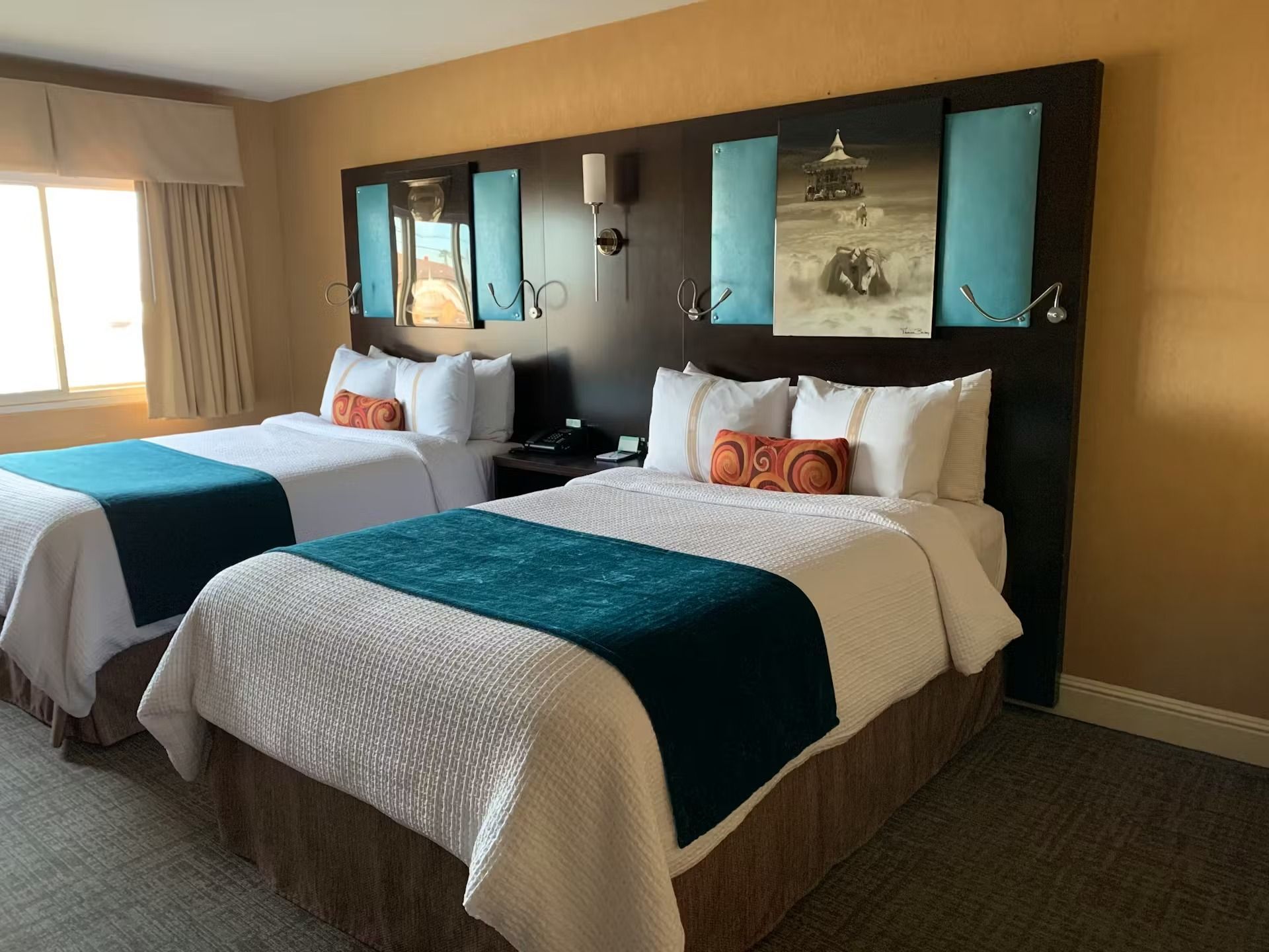 A hotel room with two beds featuring white bedding, teal runners, and a dark wood headboard with abstract wall art.