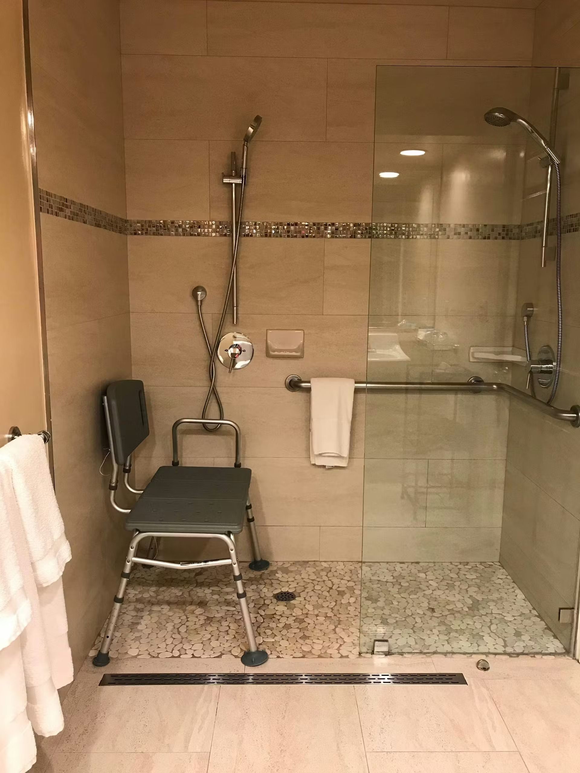 An accessible shower with a wall-mounted fold-down seat, a handheld showerhead, a grab bar, and a low-profile drain.