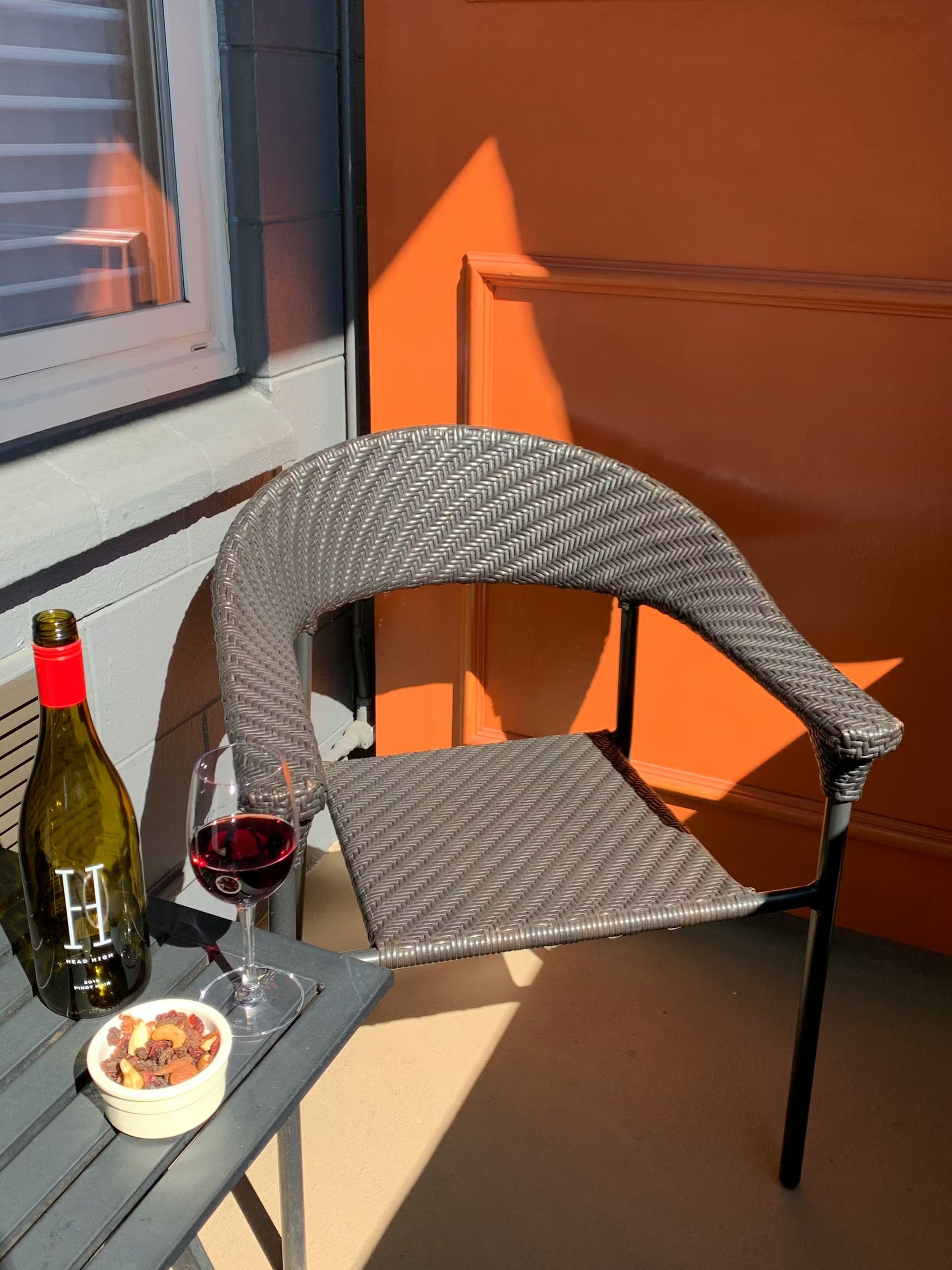 A wine bottle, a glass of red wine, and a small bowl of nuts sit on a table next to a patio chair in warm, sunny light.