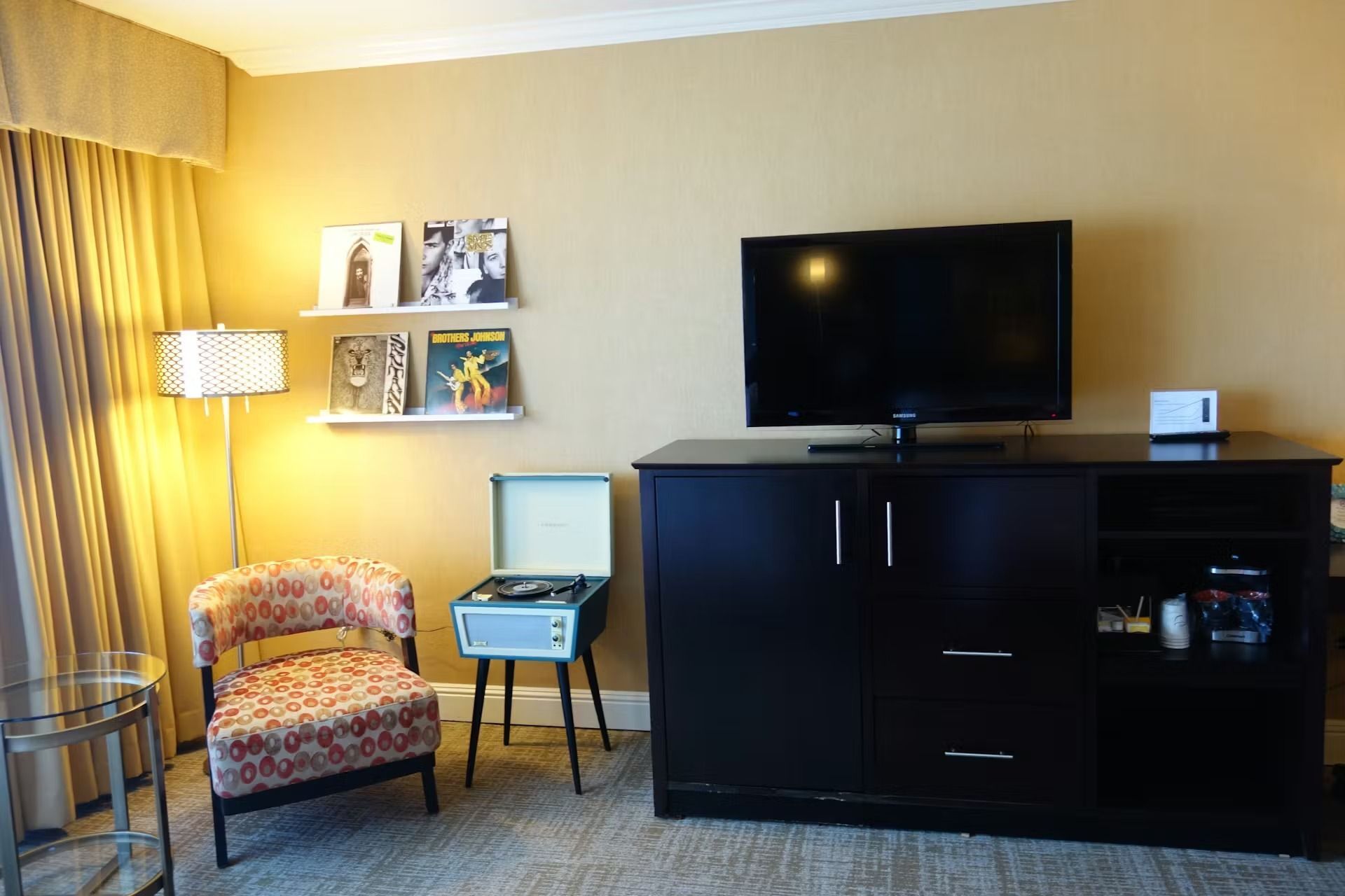 A hotel room with a television on a dark cabinet, a record player, a patterned armchair, and wall-mounted vinyl records.