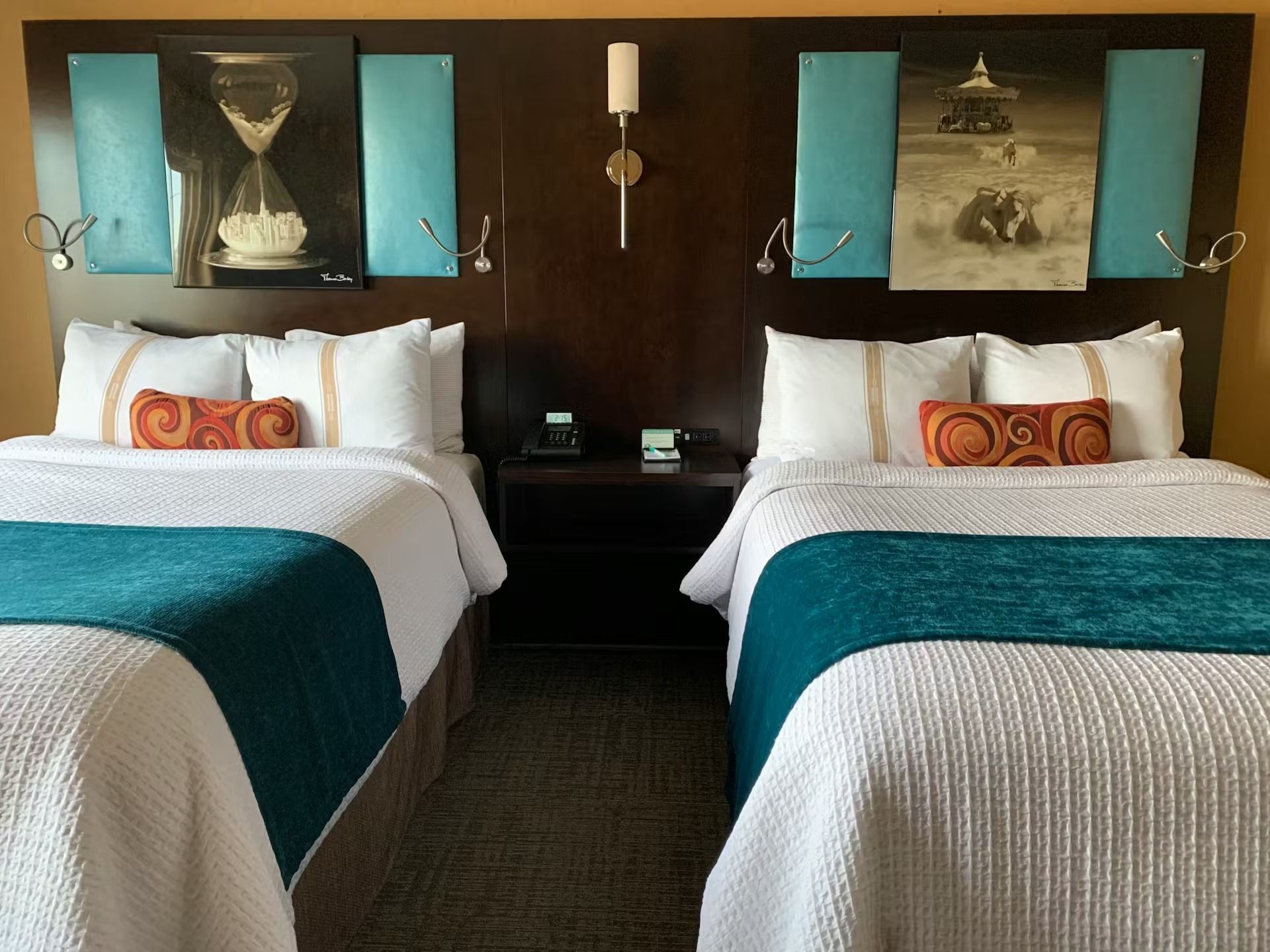 A hotel room featuring two beds with white quilts, teal runners, and orange accent pillows against a dark wood headboard.