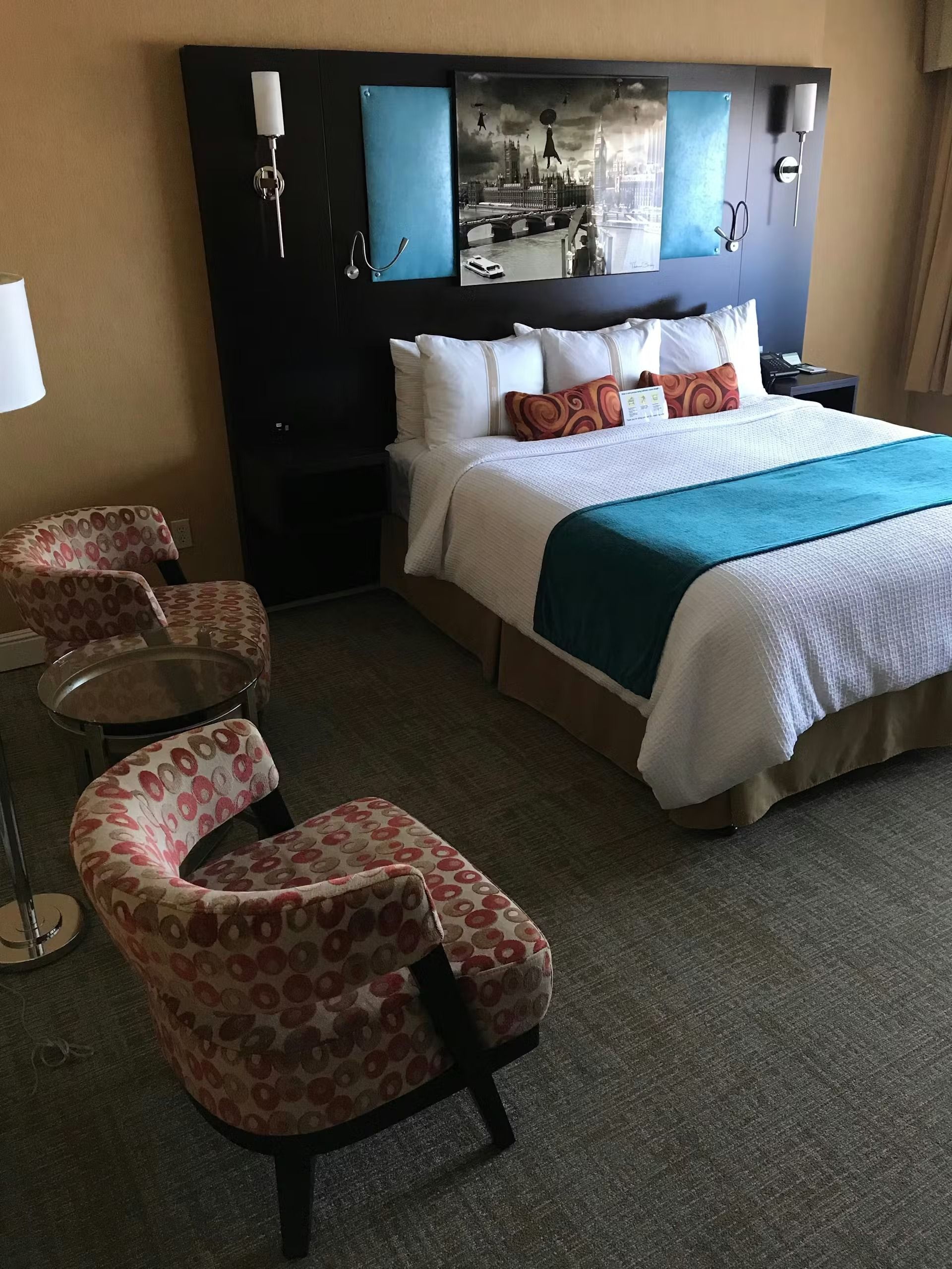 A hotel room with a large bed featuring a teal runner and patterned pillows, next to two patterned chairs and a table.