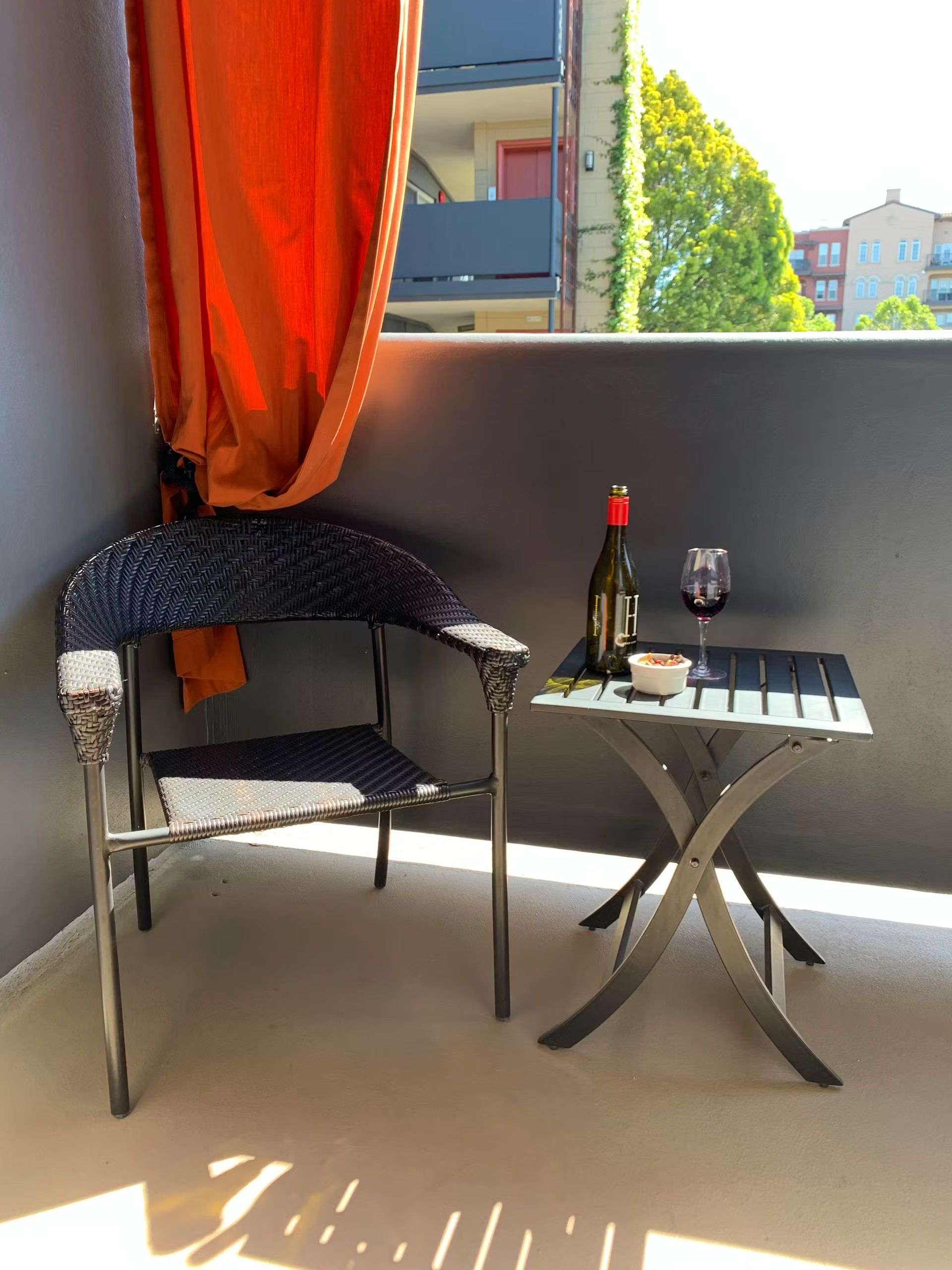A dark wicker chair and a small metal folding table with a bottle of wine and a glass sit on a sunny balcony.