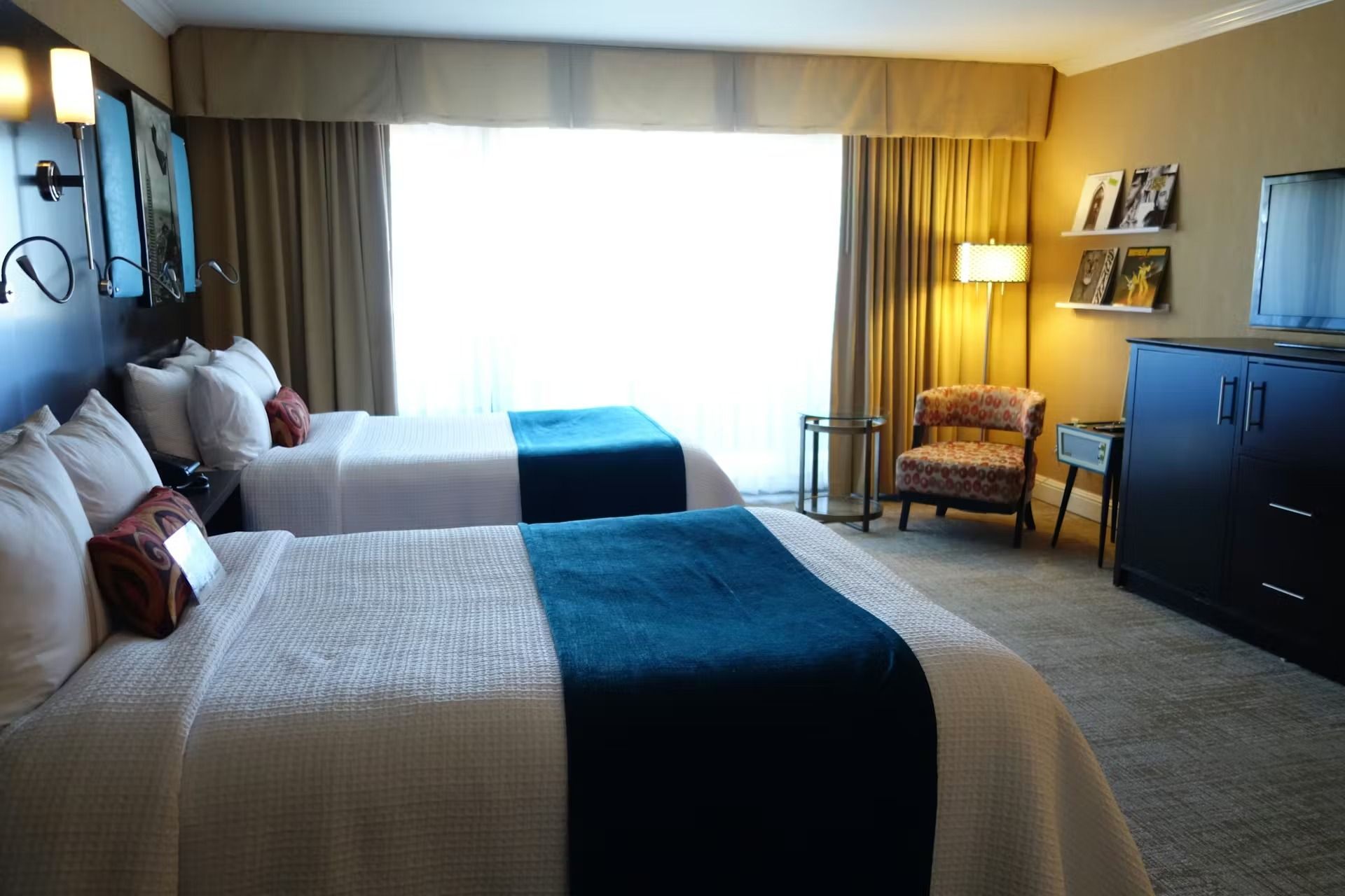 A hotel room featuring two queen beds with white linens and teal runners, an armchair, and a dark wooden dresser.