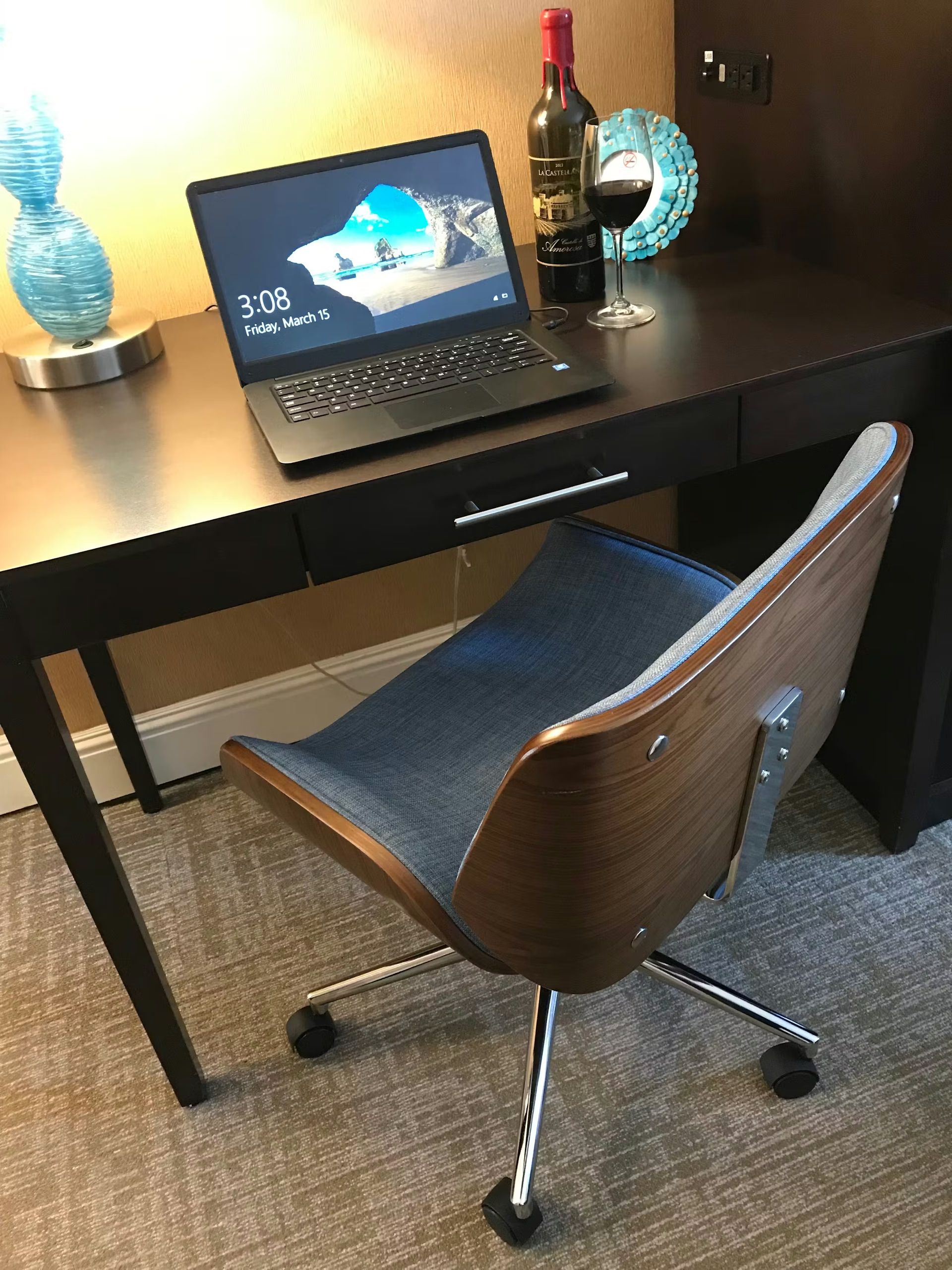 A modern desk with a laptop, wine bottle, and glass, paired with a wood-and-blue fabric rolling chair on a carpeted floor.