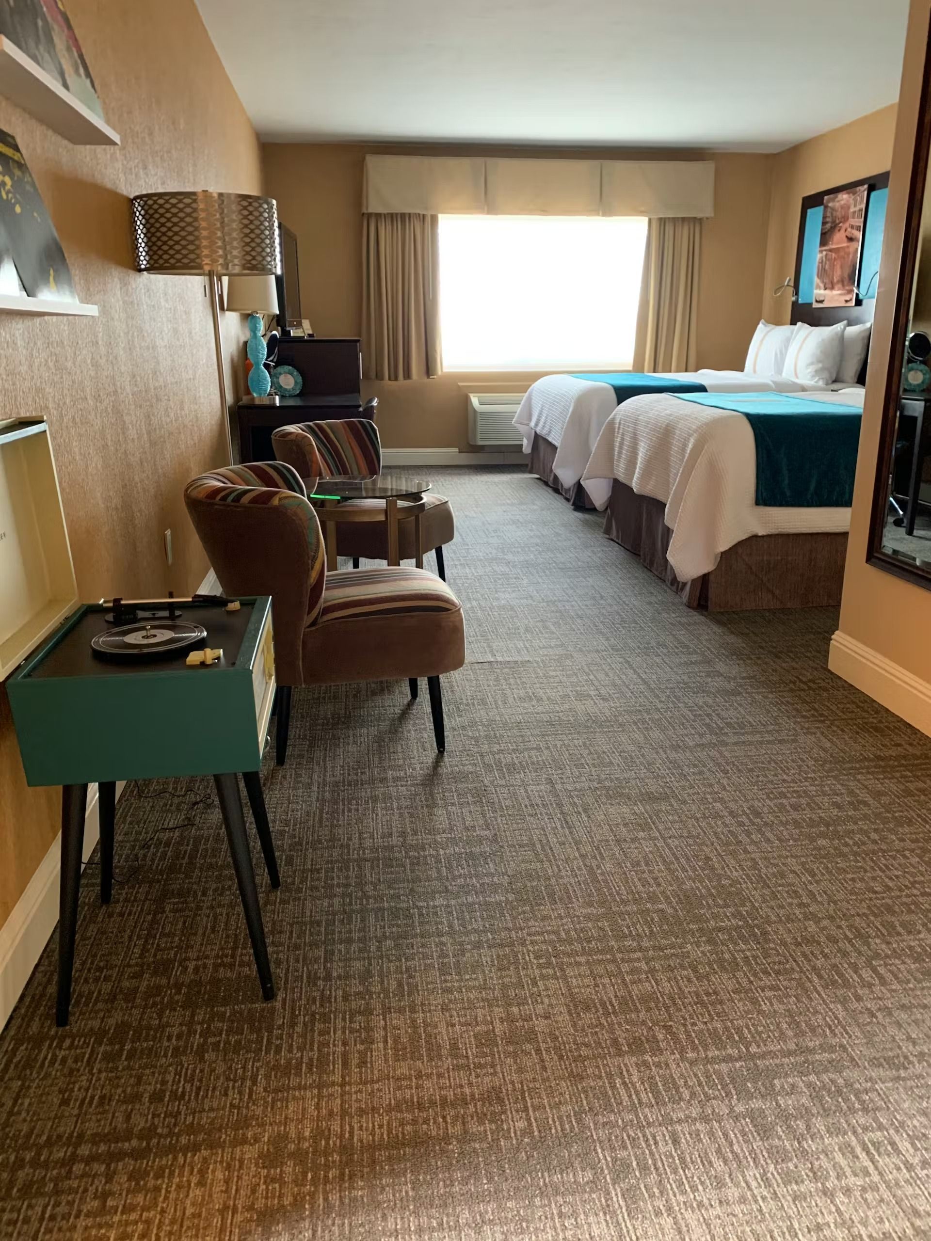 A hotel room with two beds, brown armchairs, a teal side table, and a window with light beige curtains.