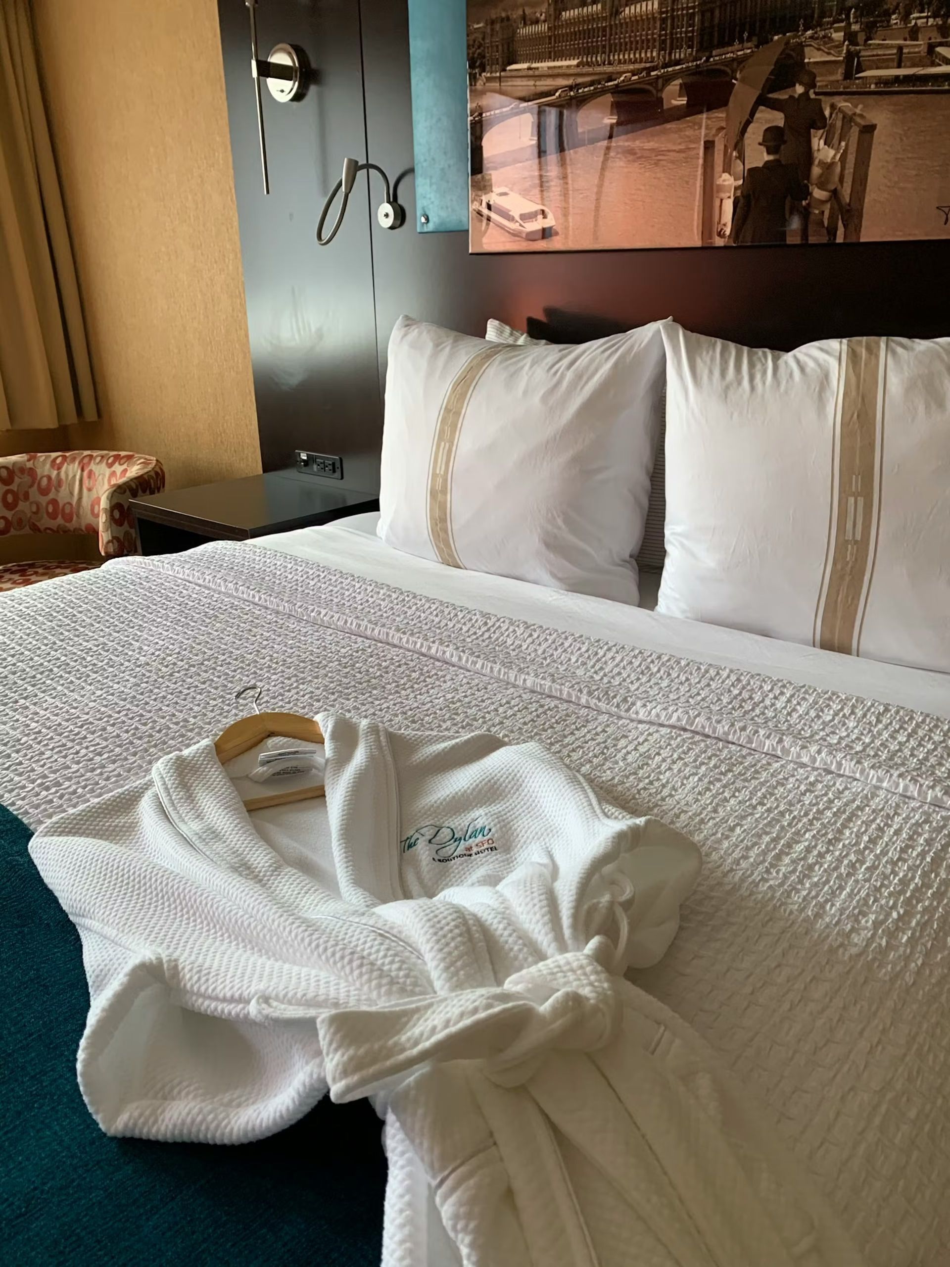 A plush white bathrobe rests on a neatly made hotel bed with two pillows and a dark headboard featuring wall art.