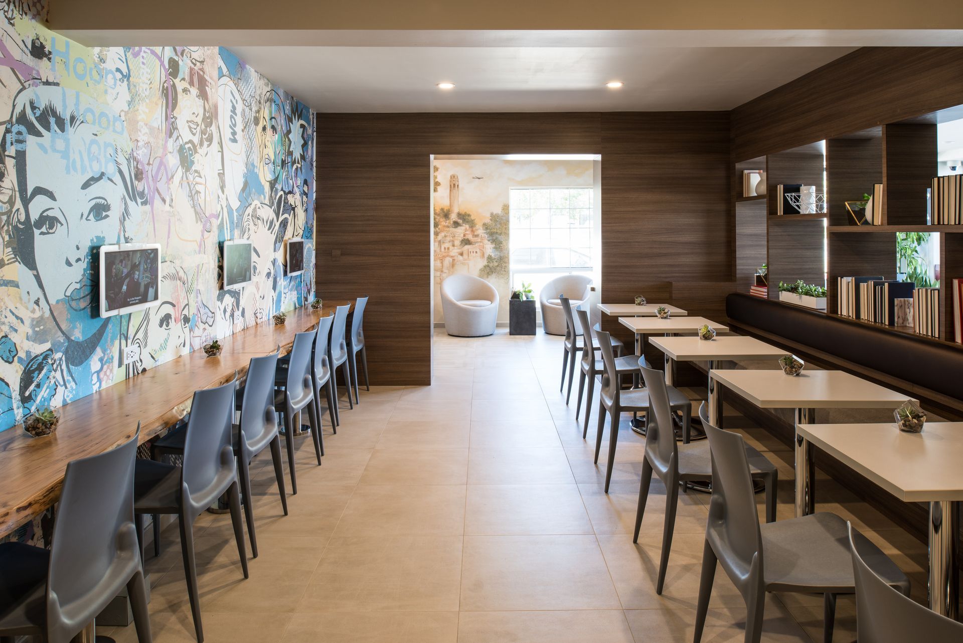 A modern communal lounge with a mural wall, long wooden desk with chairs, and rows of tables and chairs for dining.