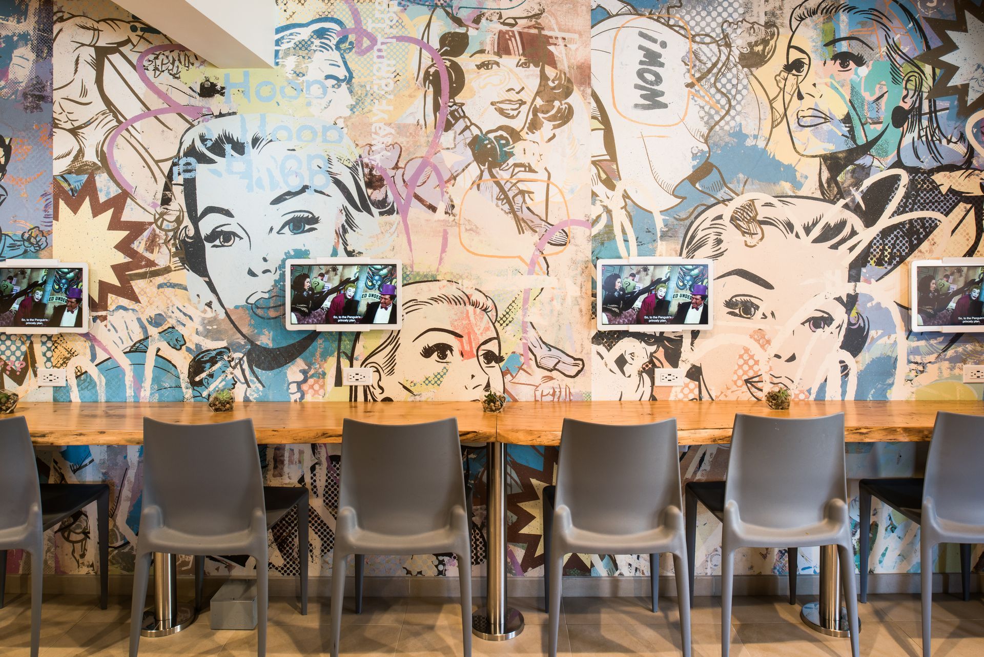 A row of gray chairs lines a wooden table against a wall decorated with colorful, pop-art style comic book illustrations.