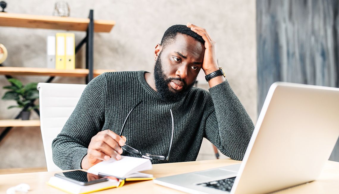 A person in a gray sweater sits at a desk with a laptop, looking stressed while holding glasses and touching their head.