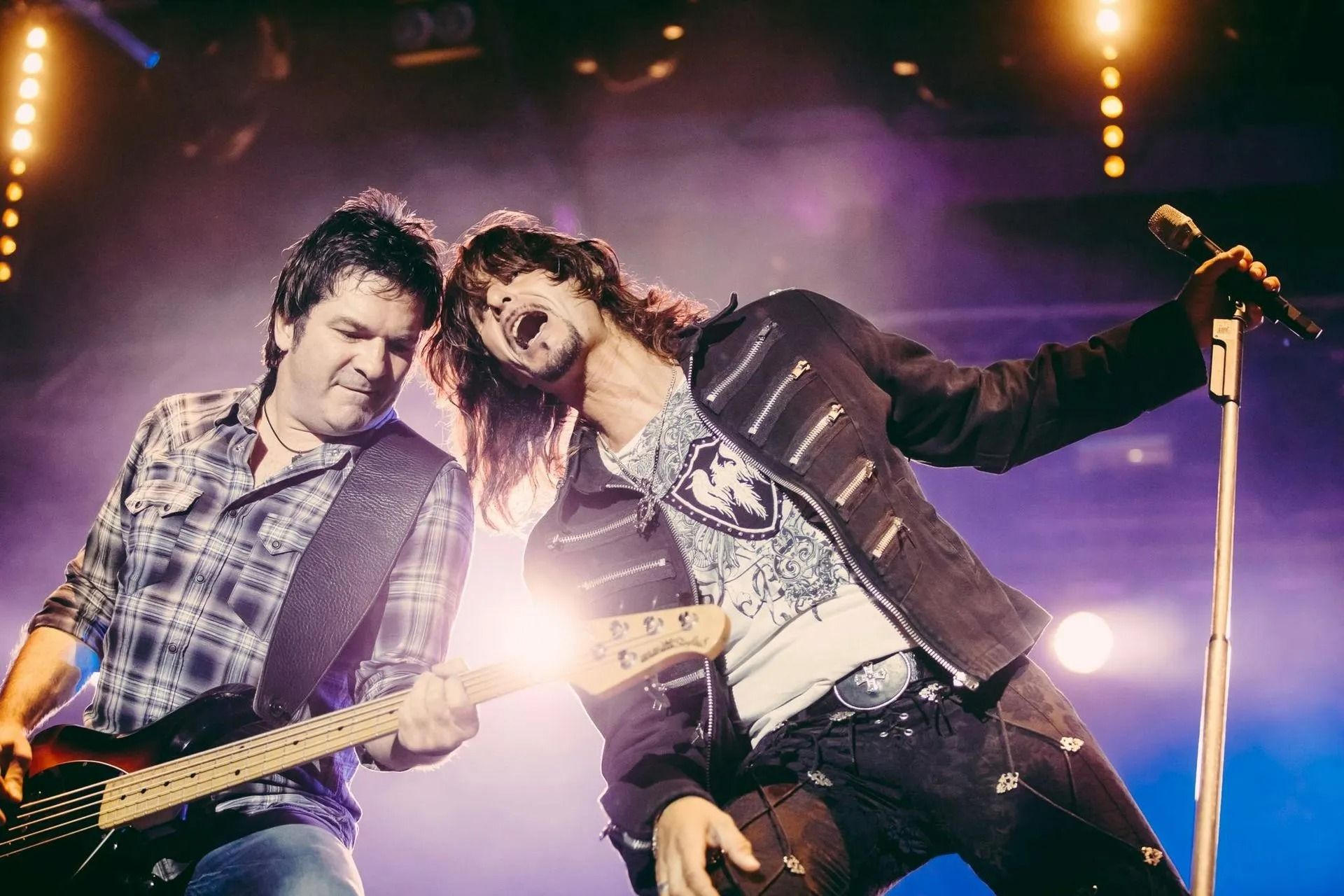 Two musicians performing on stage, one playing guitar under bright concert lights.