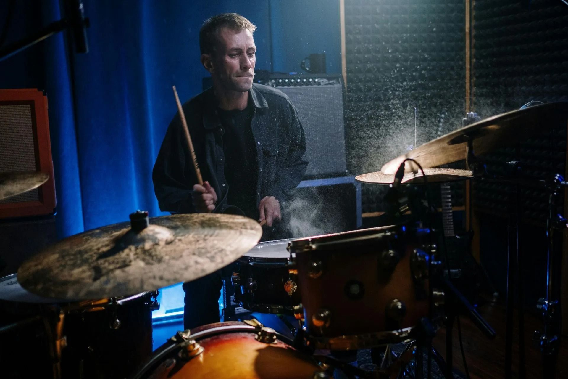 Drummer seated behind a drum kit on a dimly lit stage with blue curtains and warm spotlighting