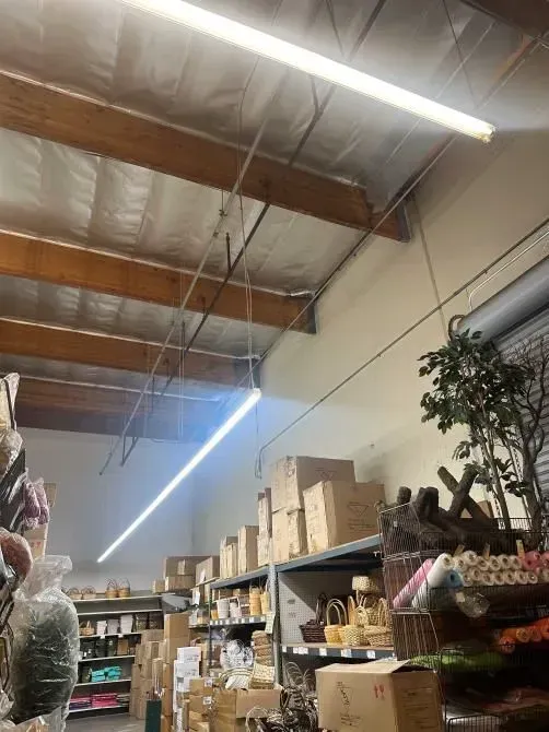 Warehouse aisle with shelves of boxes and supplies under fluorescent lights and exposed wooden beams