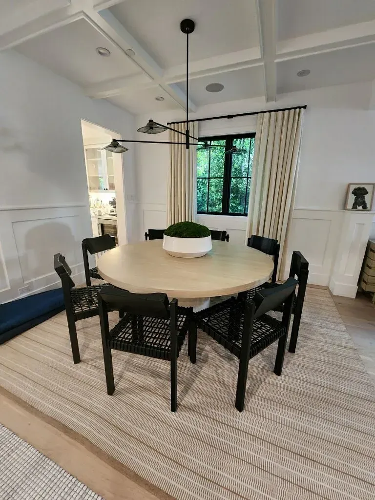 Bright dining room with round wood table, black chairs, striped rug, and large window with curtains